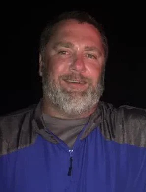 A man with a beard smiling, wearing a black and blue jacket, against a dark background.