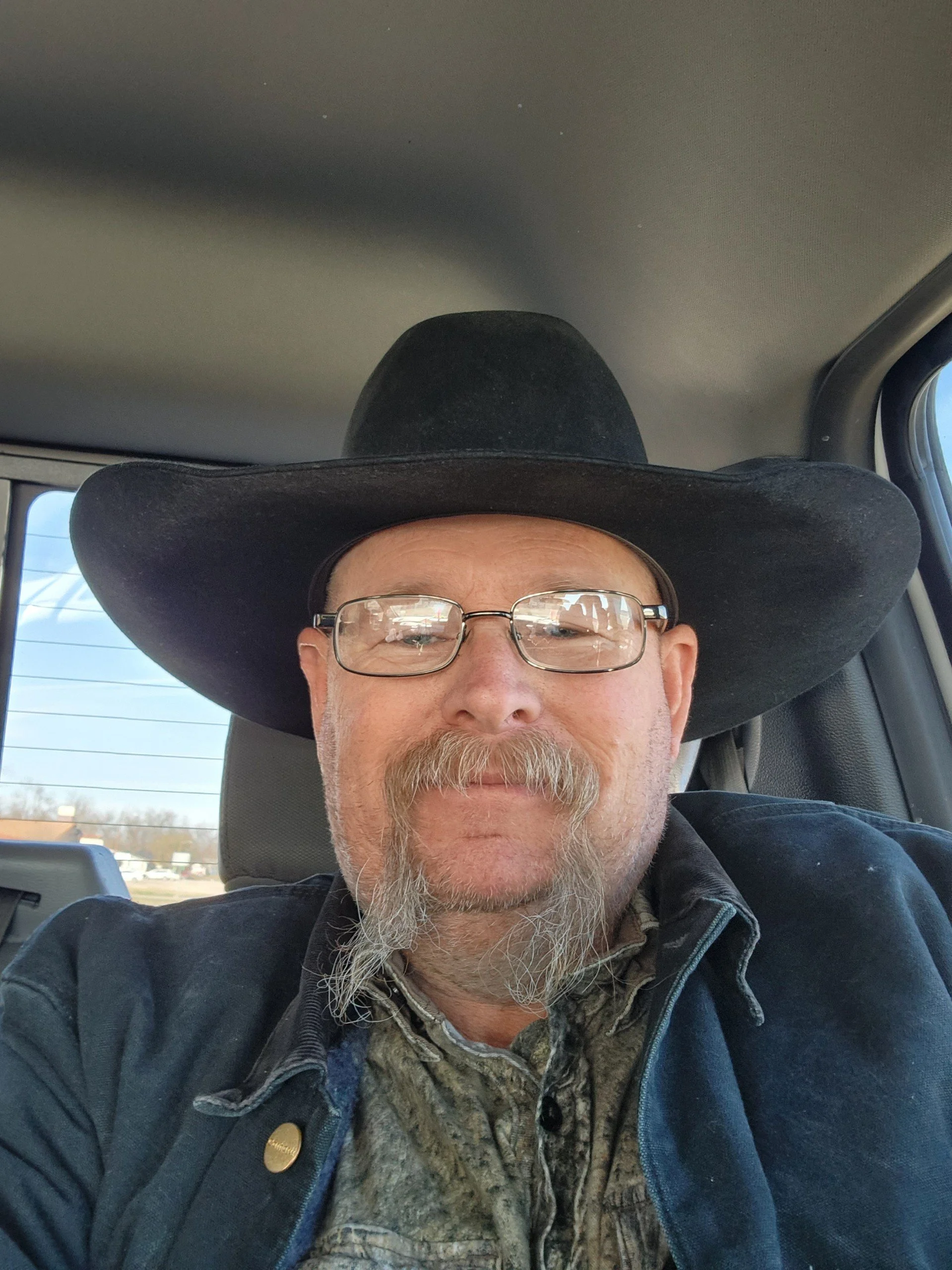 A man with a beard and mustache wearing glasses, a black cowboy hat, and a denim jacket taking a selfie inside a vehicle.