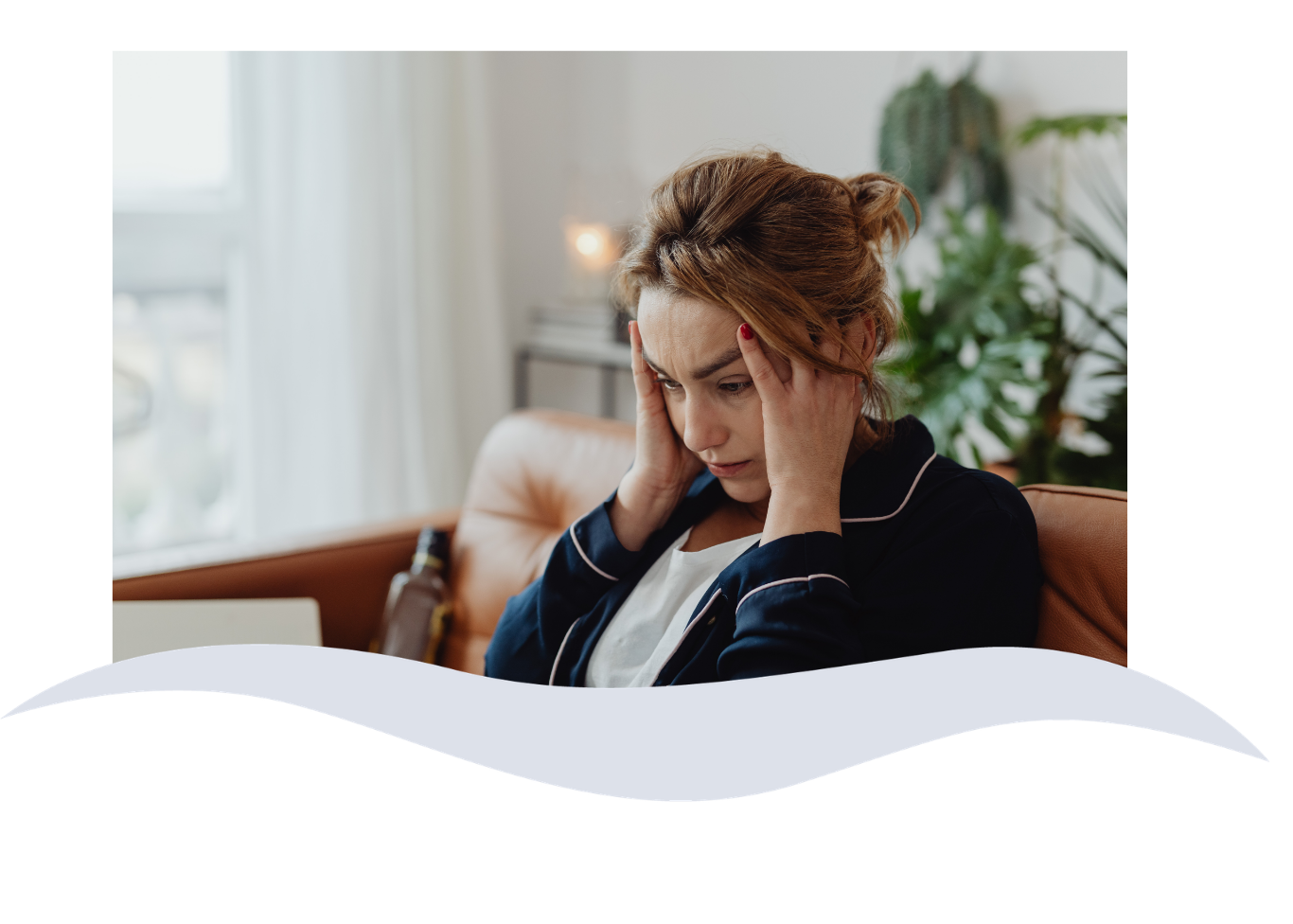 A woman sitting on a couch holding her head in distress, with a park or outdoor view visible through the window in the background.