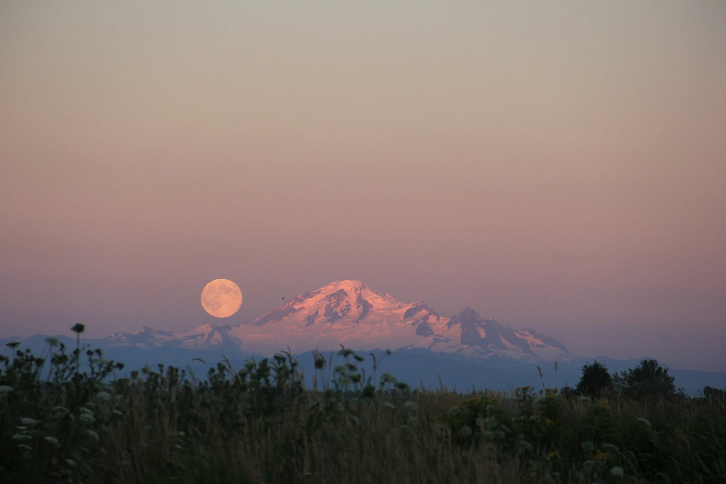 A mountain with snow-covered peak at sunset, with the moon visible in the sky above the mountain.