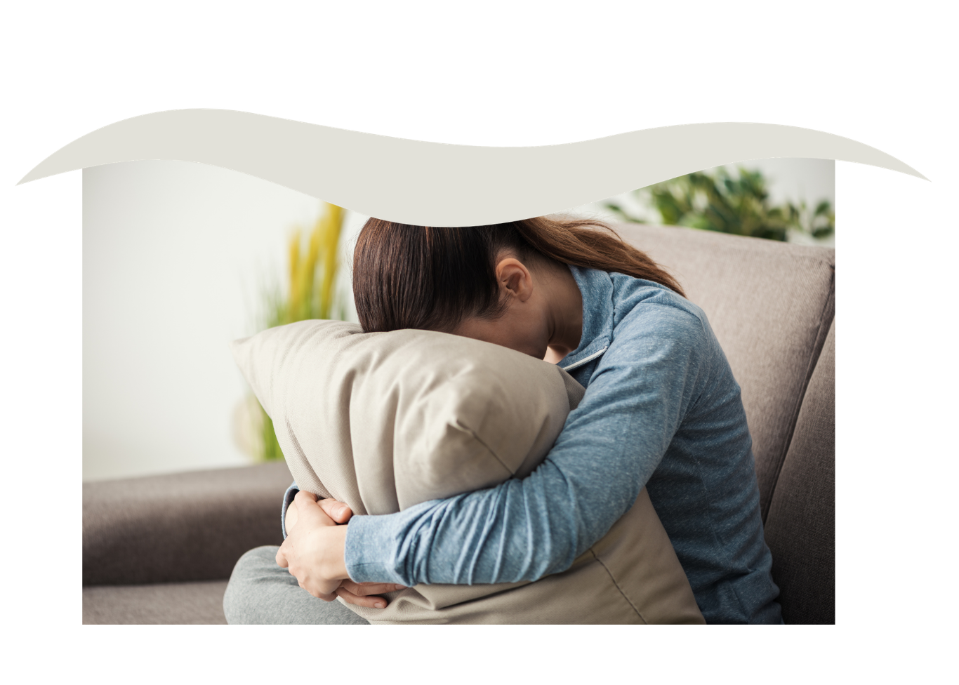 A woman with brown hair wearing a blue long-sleeve shirt sitting on a couch, hugging a beige pillow and resting her head on it with her eyes closed.