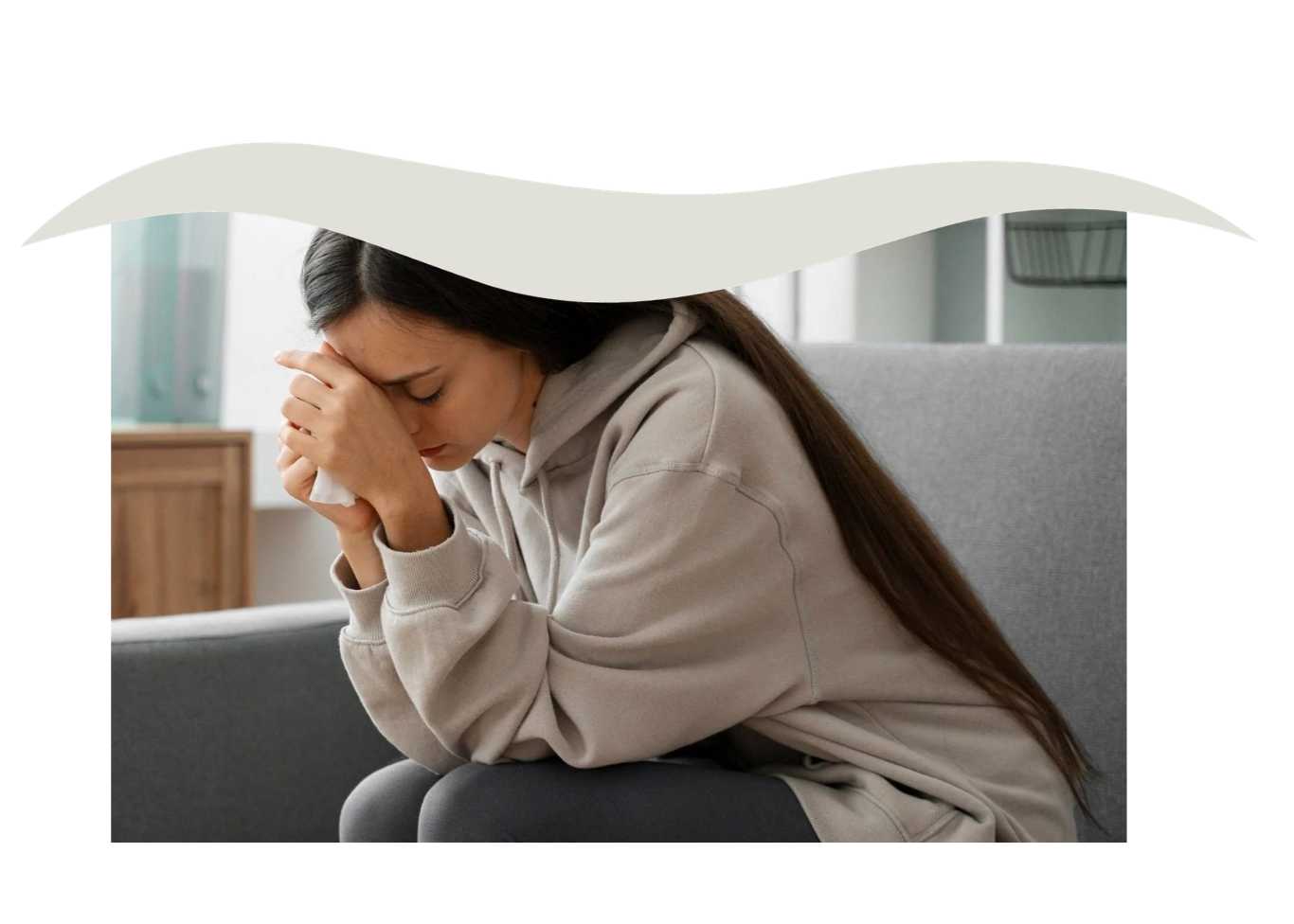 A young woman sitting on a couch, holding a tissue to her face, appearing upset or distressed.