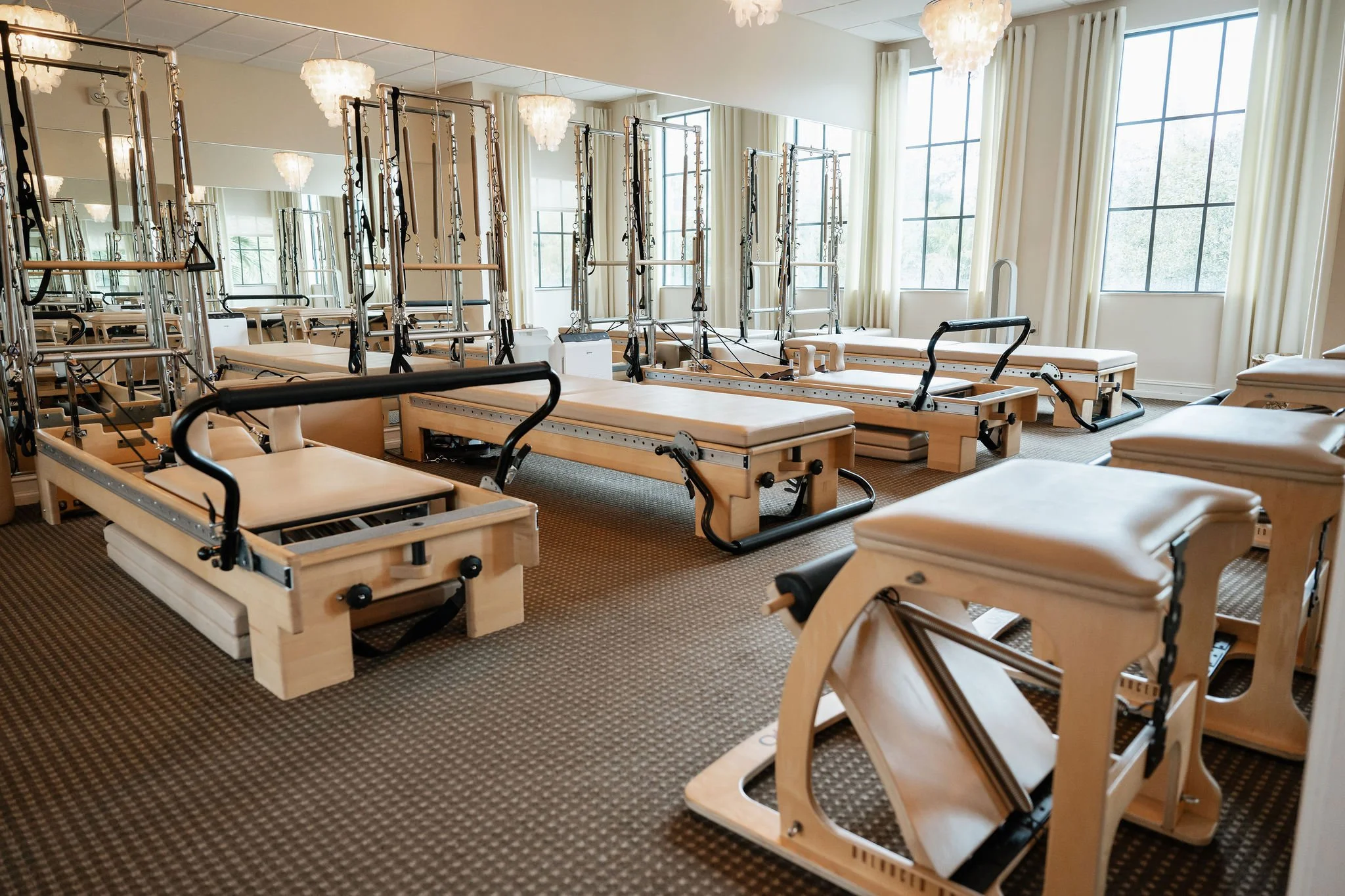 Pilates reformers and chairs in a well-lit, spacious studio with large windows and elegant chandeliers. Group class room at Core Elements in Jupiter, FL