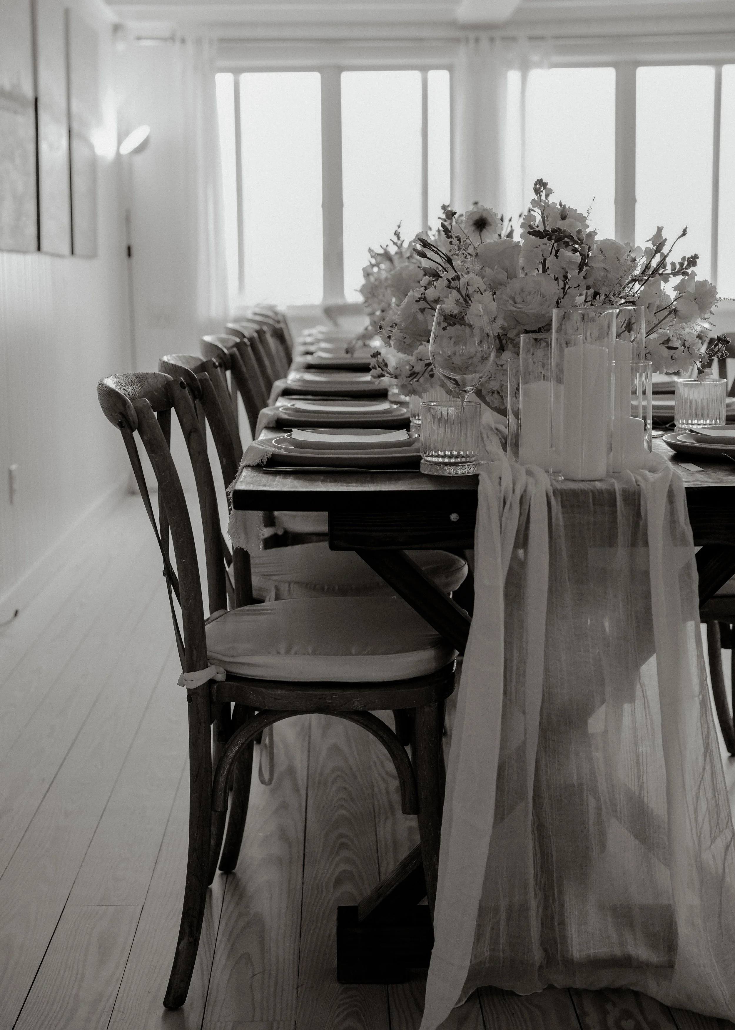 A dining room table set with white plates, glasses, and a large floral centerpiece in a glass vase, with a light-colored tablecloth and chairs lined up along one side. Sunlight streams through large windows in the background.