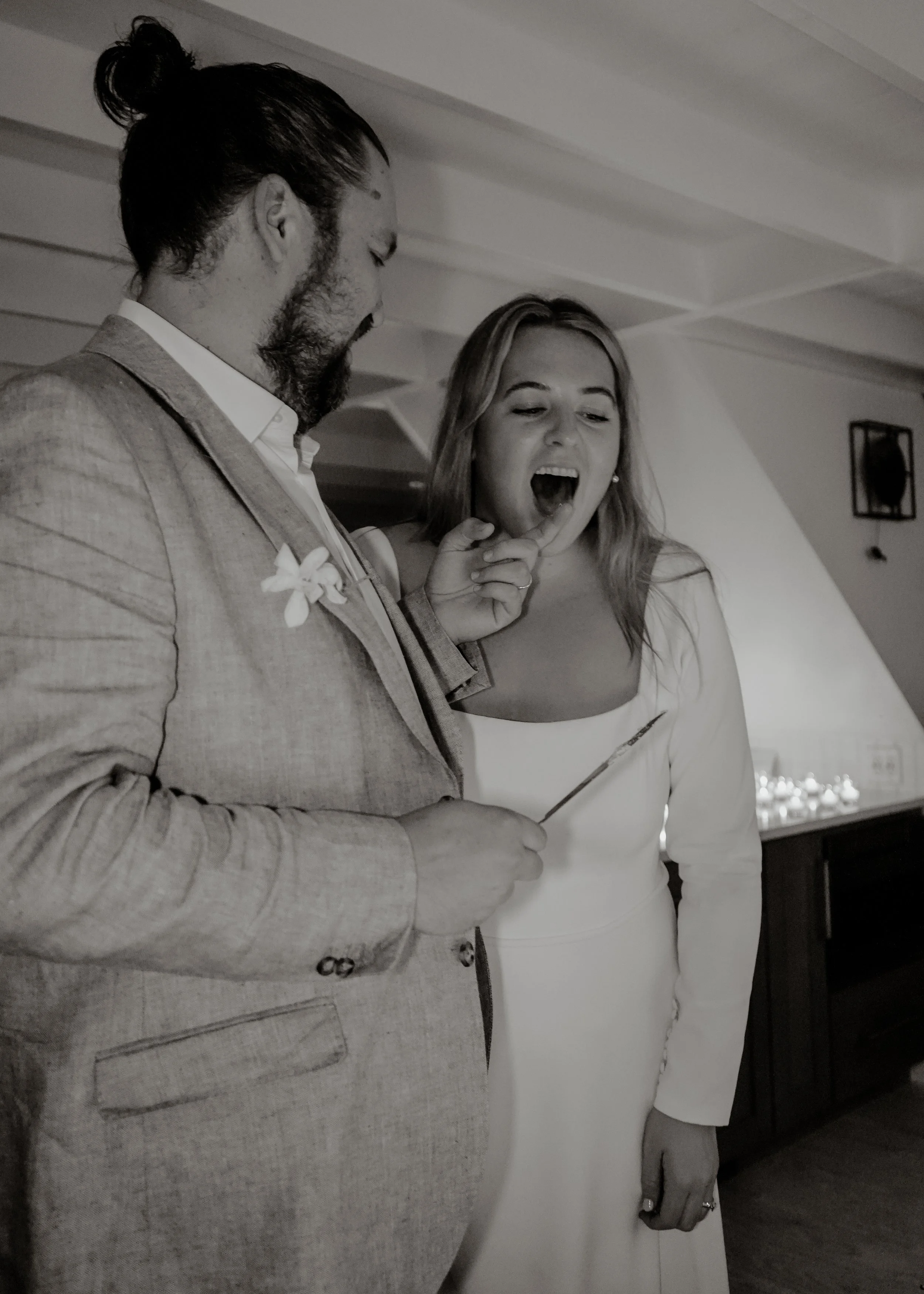 A man and woman stand close together indoors, with the woman opening her mouth and holding up her index finger near her face, while the man looks down. The woman is wearing a light-colored dress, and the man is dressed in a light-colored blazer with 