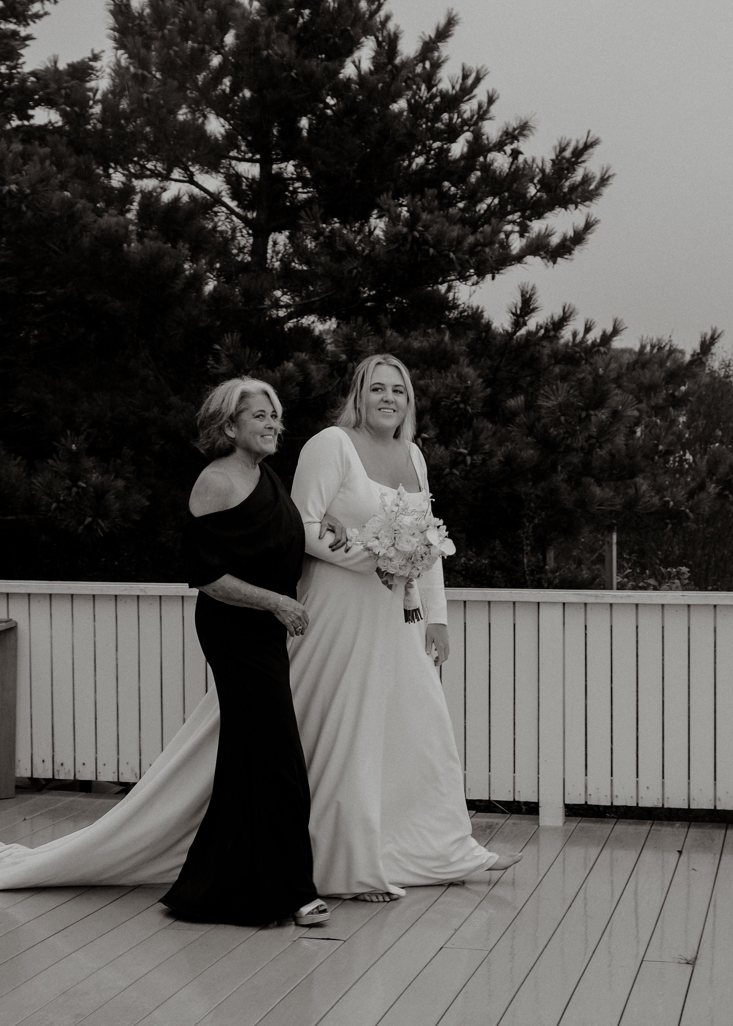 A woman in a wedding dress holding a bouquet, with an older woman beside her, both standing outdoors in front of a pine tree, during a wedding or similar celebration.