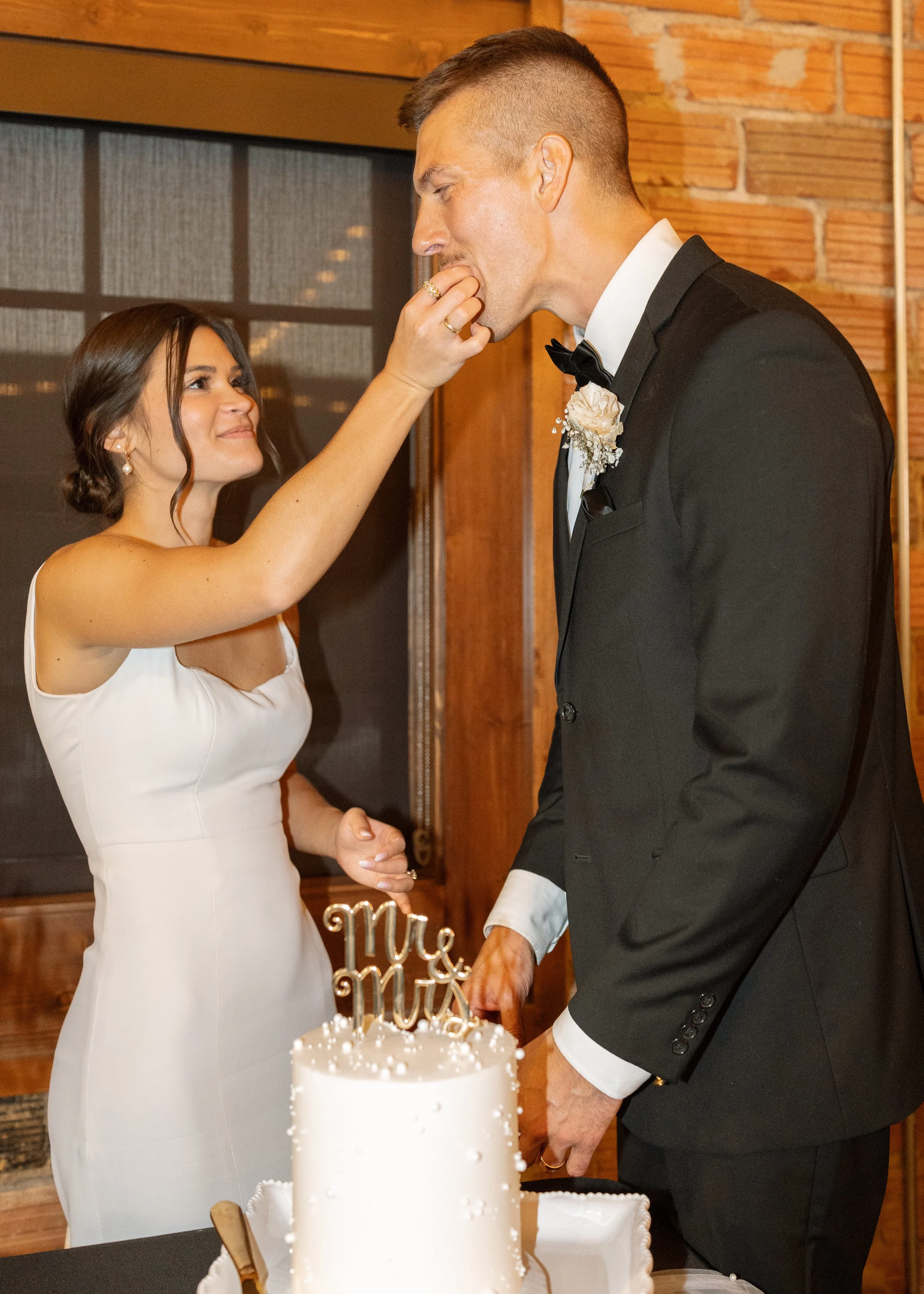 A bride and groom stand together at a wedding reception. The bride is smiling and looks happy, wearing a white dress. The groom is dressed in a black tuxedo with a white shirt and bow tie, and is gently holding the bride's hand. In the foreground, th