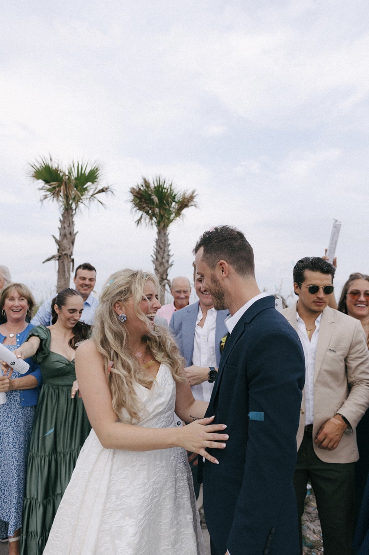 A bride and groom smiling and looking at each other at their outdoor wedding ceremony, surrounded by friends and family with palm trees in the background.