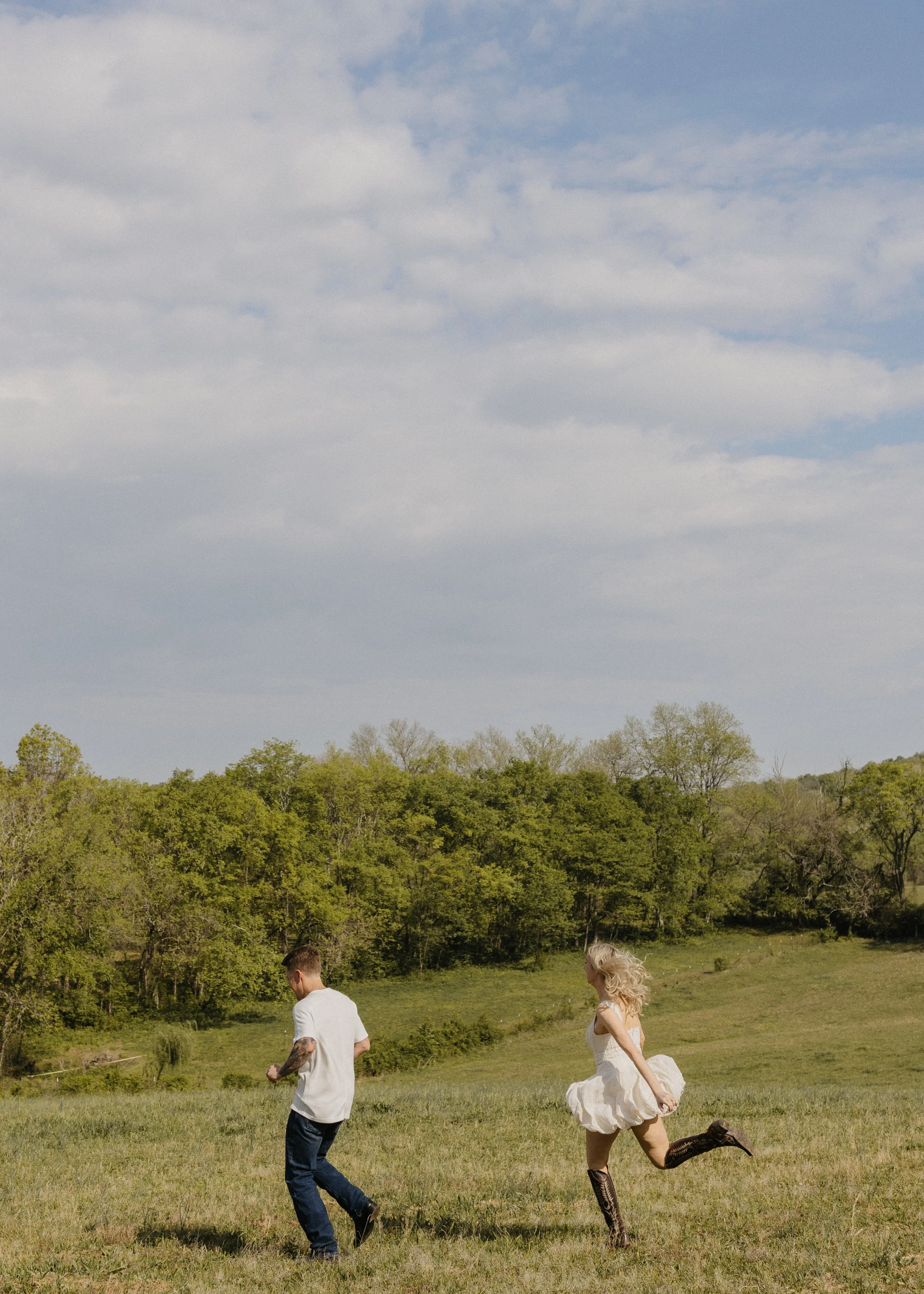 A man in a white shirt and jeans, and a woman in a white dress with cowboy boots, running on a grassy field during daytime under a blue sky with clouds, surrounded by green trees.