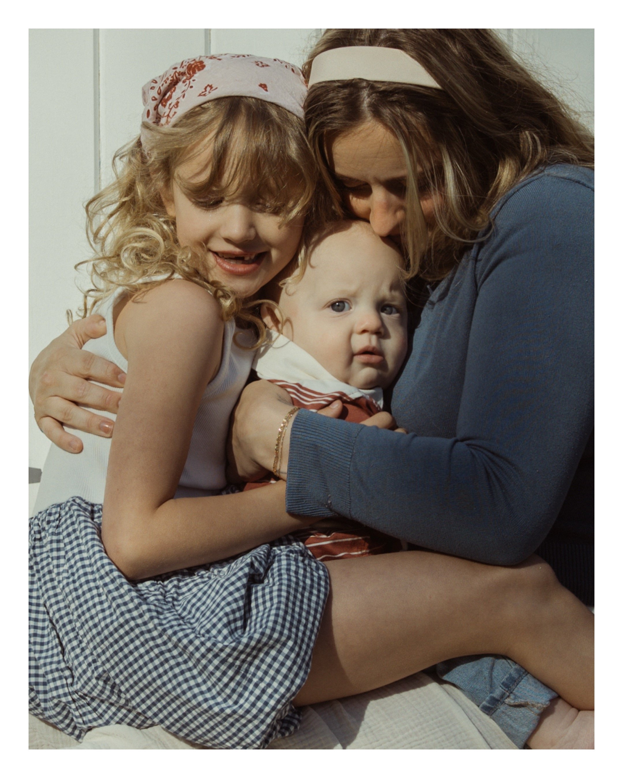 A young woman with long hair and a young girl with curly blonde hair hugging and smiling, with a baby in front of them showing a serious expression. They are sitting together on a white quilted surface. The woman is wearing a blue top and the girl a 