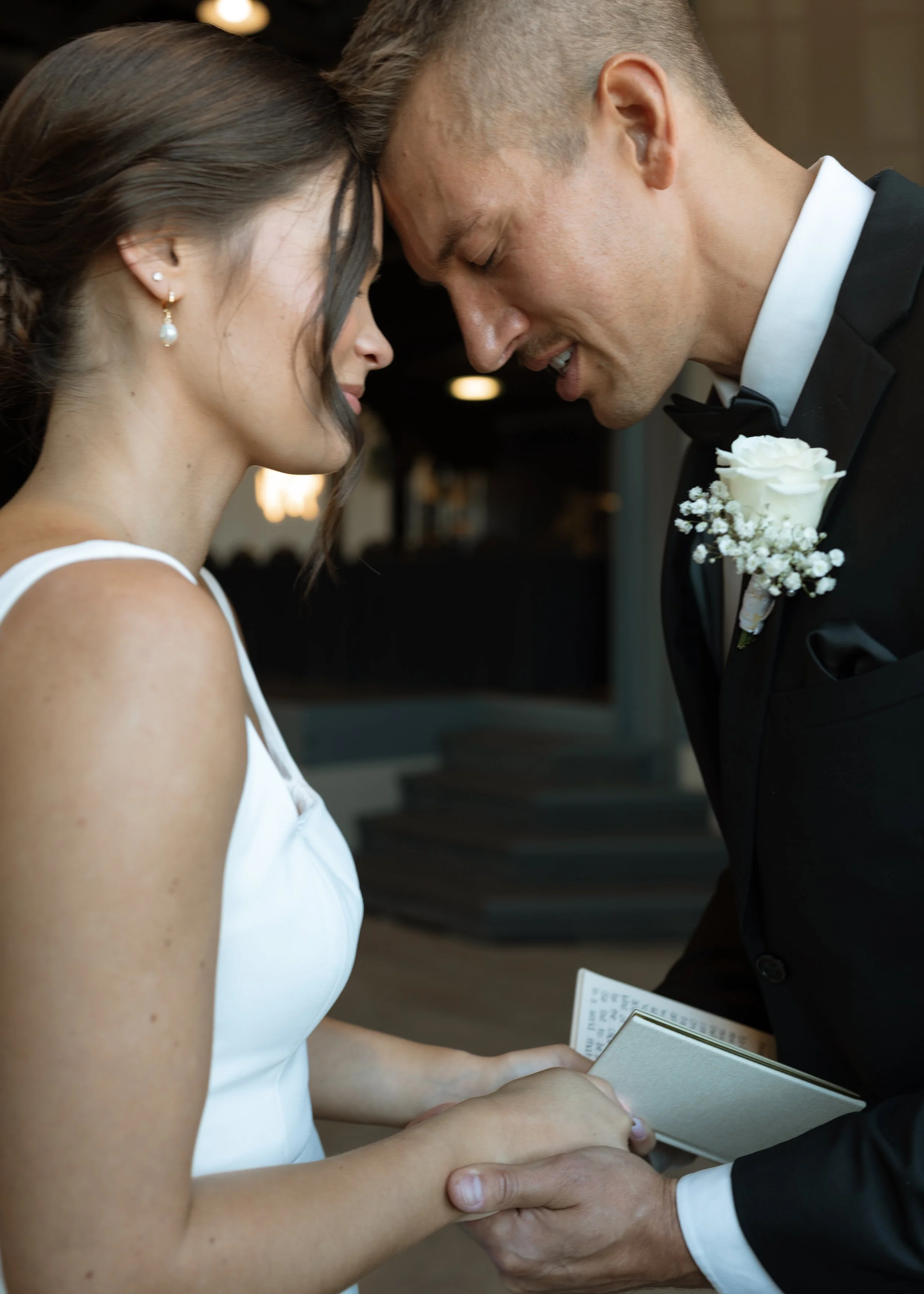 A bride and groom with their foreheads touching, holding hands during their wedding ceremony. The groom is wearing a tuxedo with a white rose boutonniere, and the bride is in a white dress with pearl earrings. They are smiling with their eyes closed.