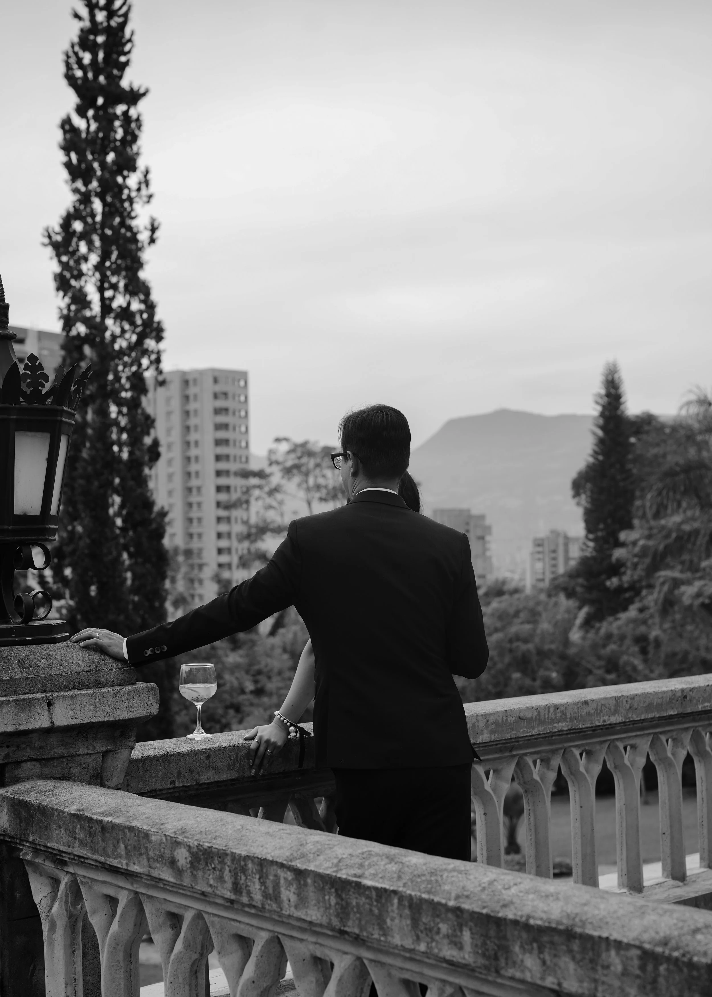 A black and white photo of a man in a suit leaning on a stone balcony with a glass of wine placed on the ledge, overlooking an urban landscape with trees and buildings in the background.