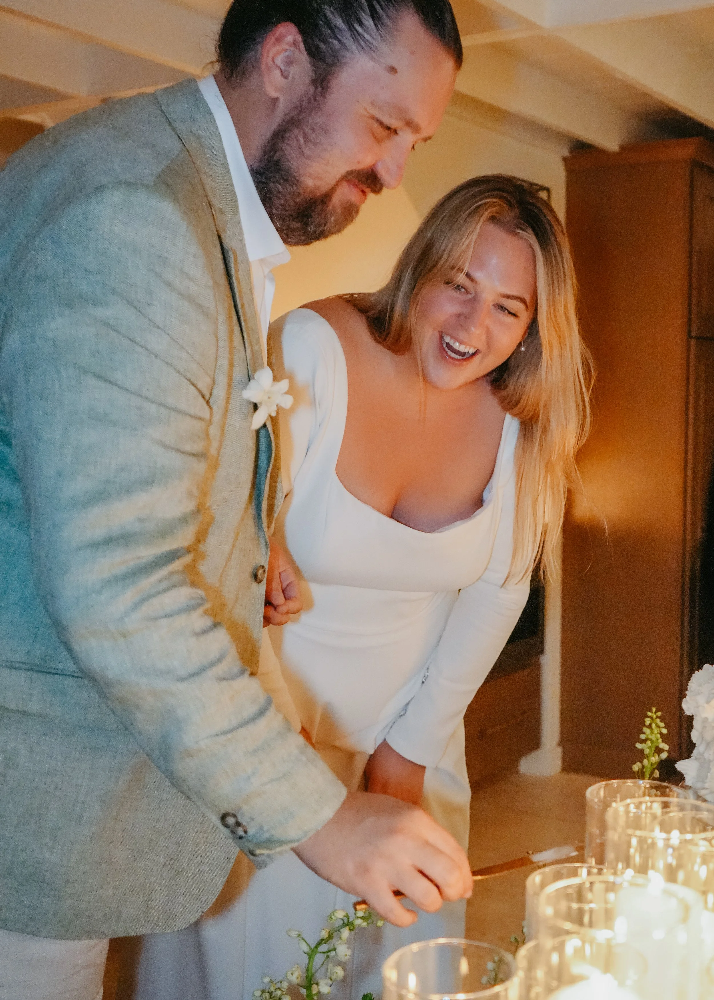 A man in a beige suit and white shirt is standing next to a woman in a white dress, both smiling and looking down at a table with candles and flowers, possibly during a celebration or ceremony.