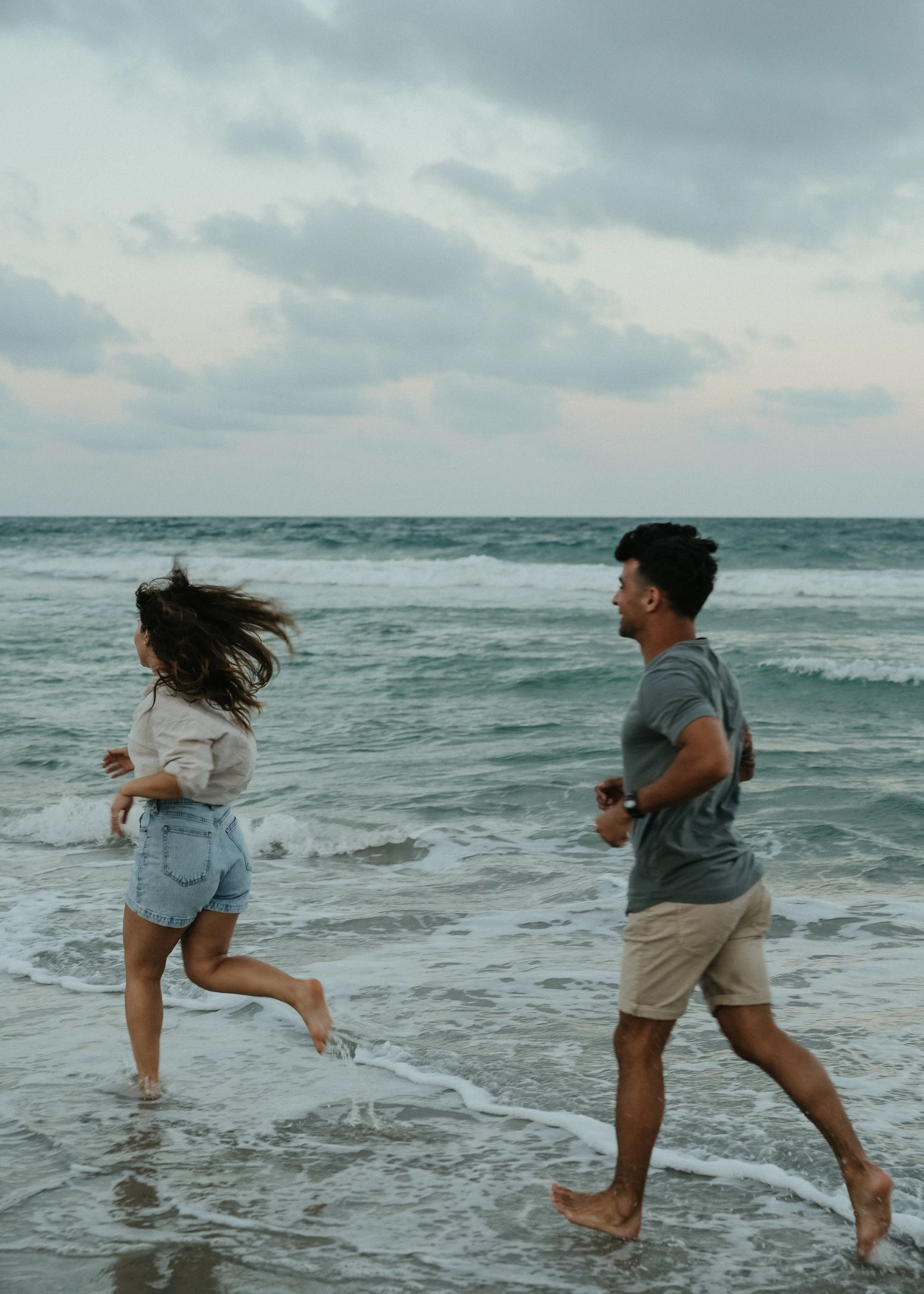 Two people, a woman and a man, are running along the shore of a beach with waves crashing around their feet. The woman has long, windblown hair and is wearing denim shorts and a light-colored top. The man has short, dark hair and is wearing a gray T-shirt and khaki shorts. The sky is cloudy and overcast.