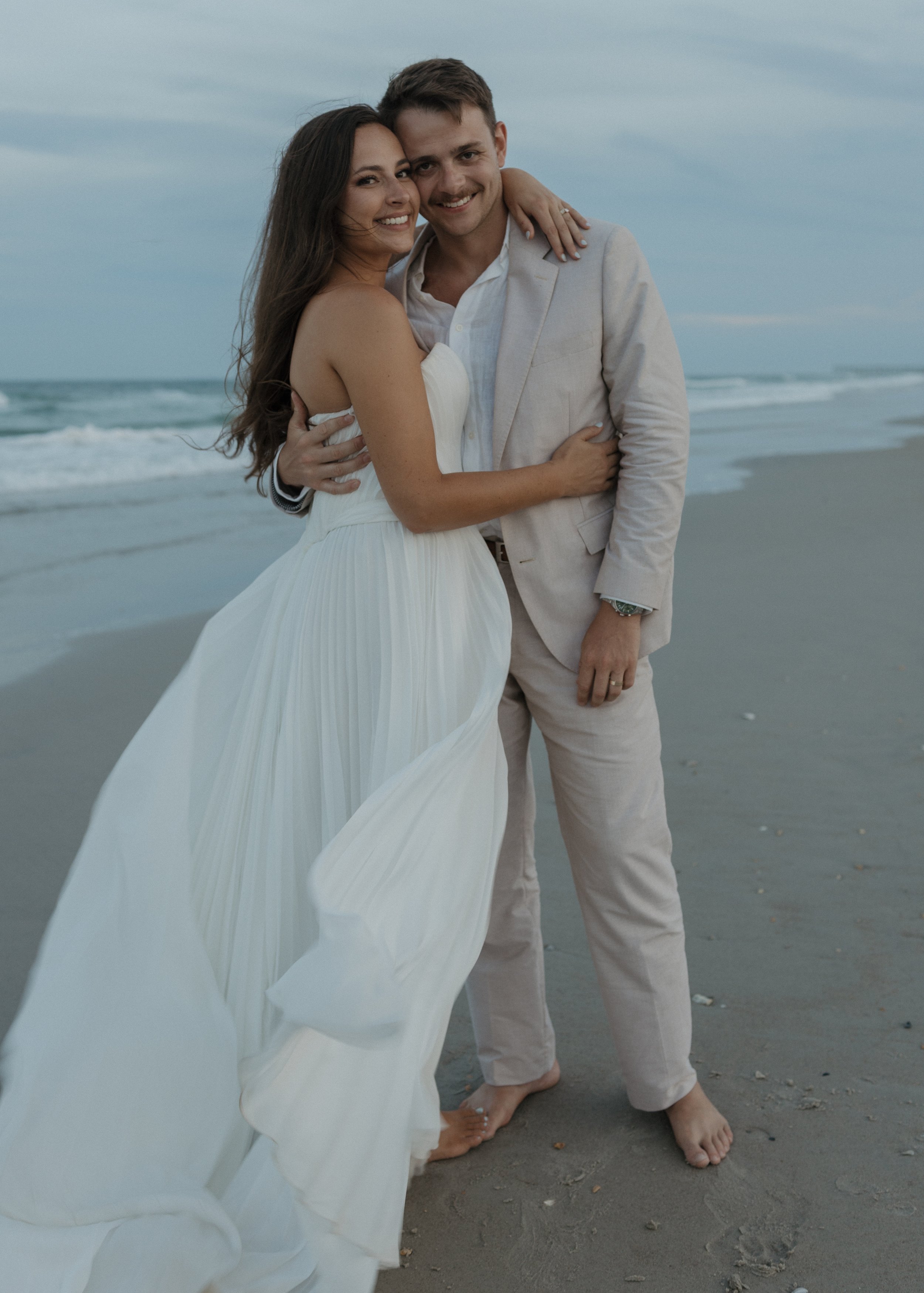 A smiling couple embracing on a beach, with the woman wearing a white dress and the man in a light-colored suit, both barefoot on the sand.