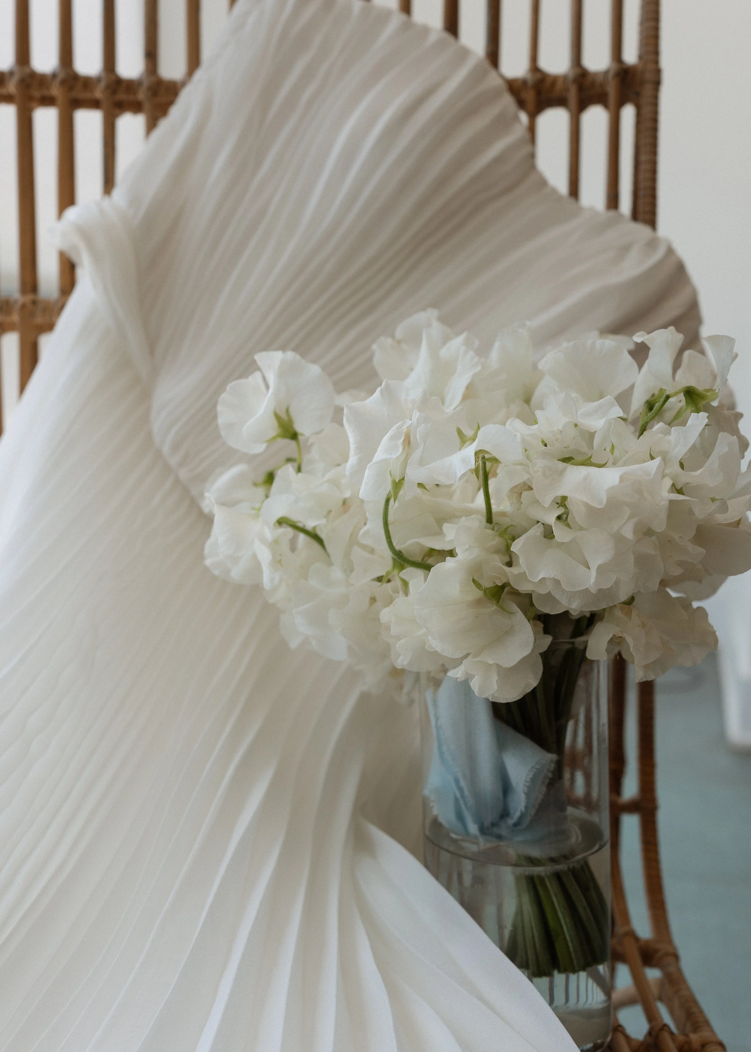 Close-up of white orchids in a glass vase against a rattan headboard with a white decorative fan in the background.