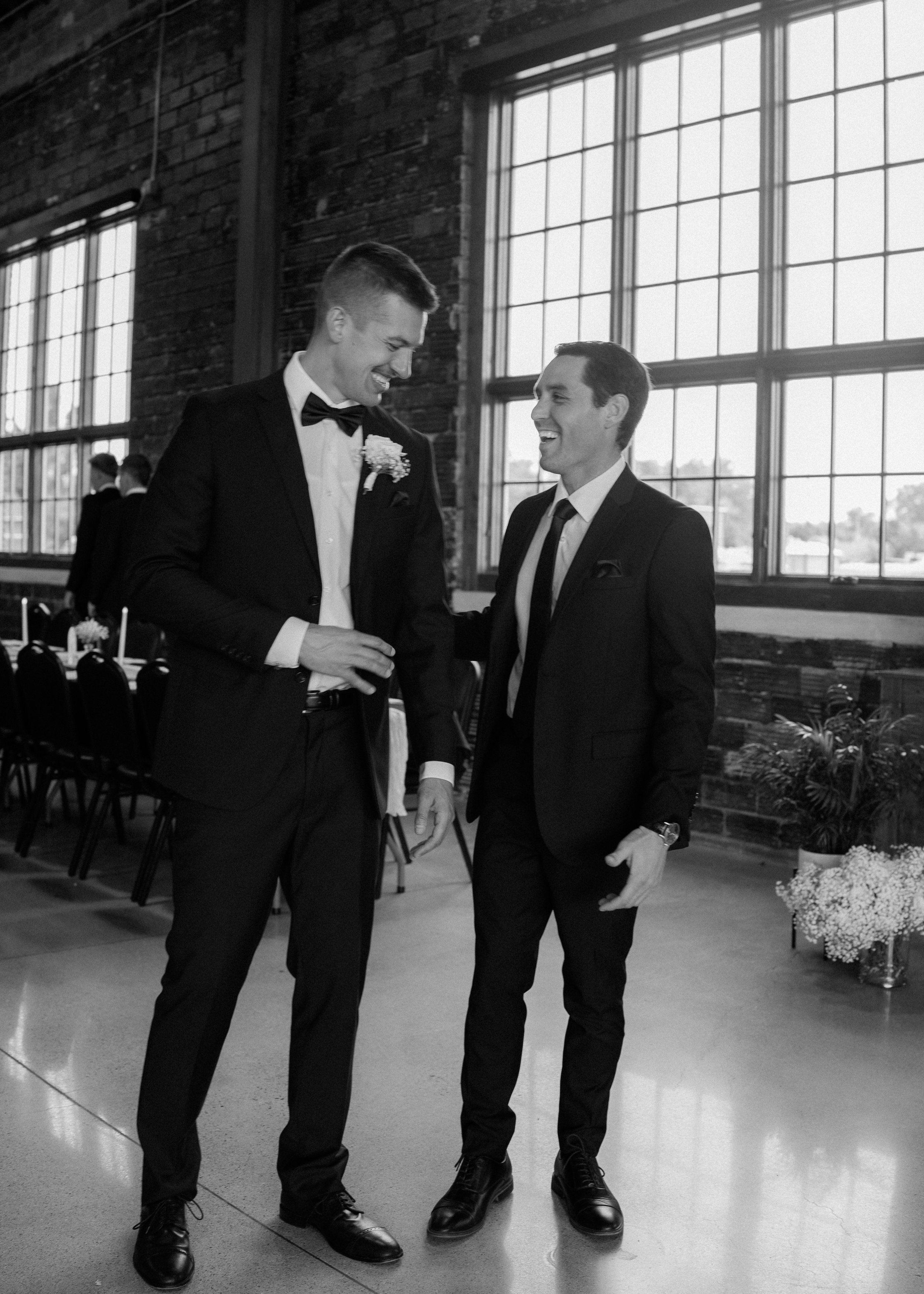 Two men in tuxedos laughing and talking in a large industrial-style room with big windows, exposed brick walls, and folding chairs in the background.