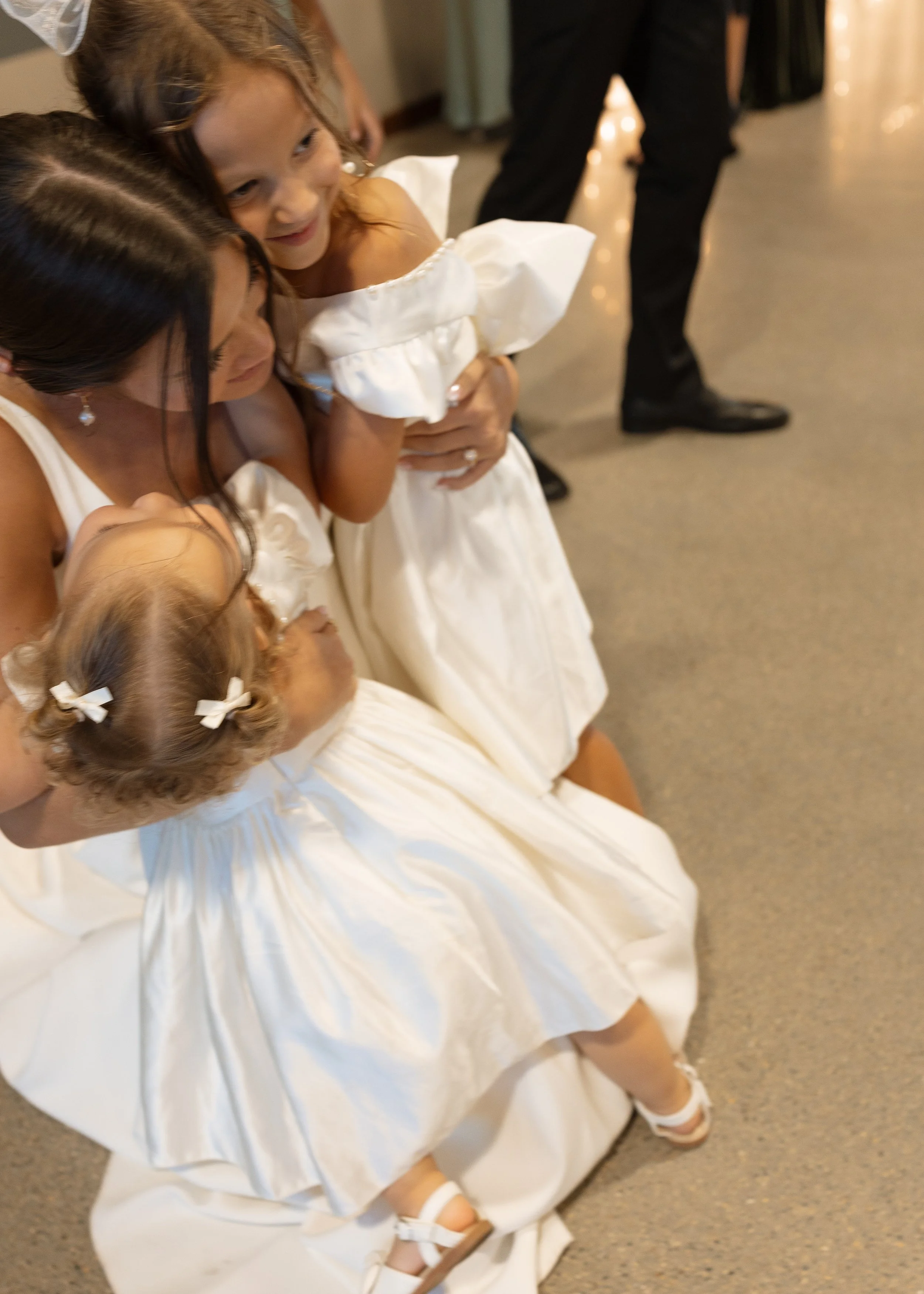 Two young girls in white dresses and white shoes, one with dark hair in pigtails with white bows and the other with light brown hair, are hugging each other and smiling at each other indoors at a formal event.