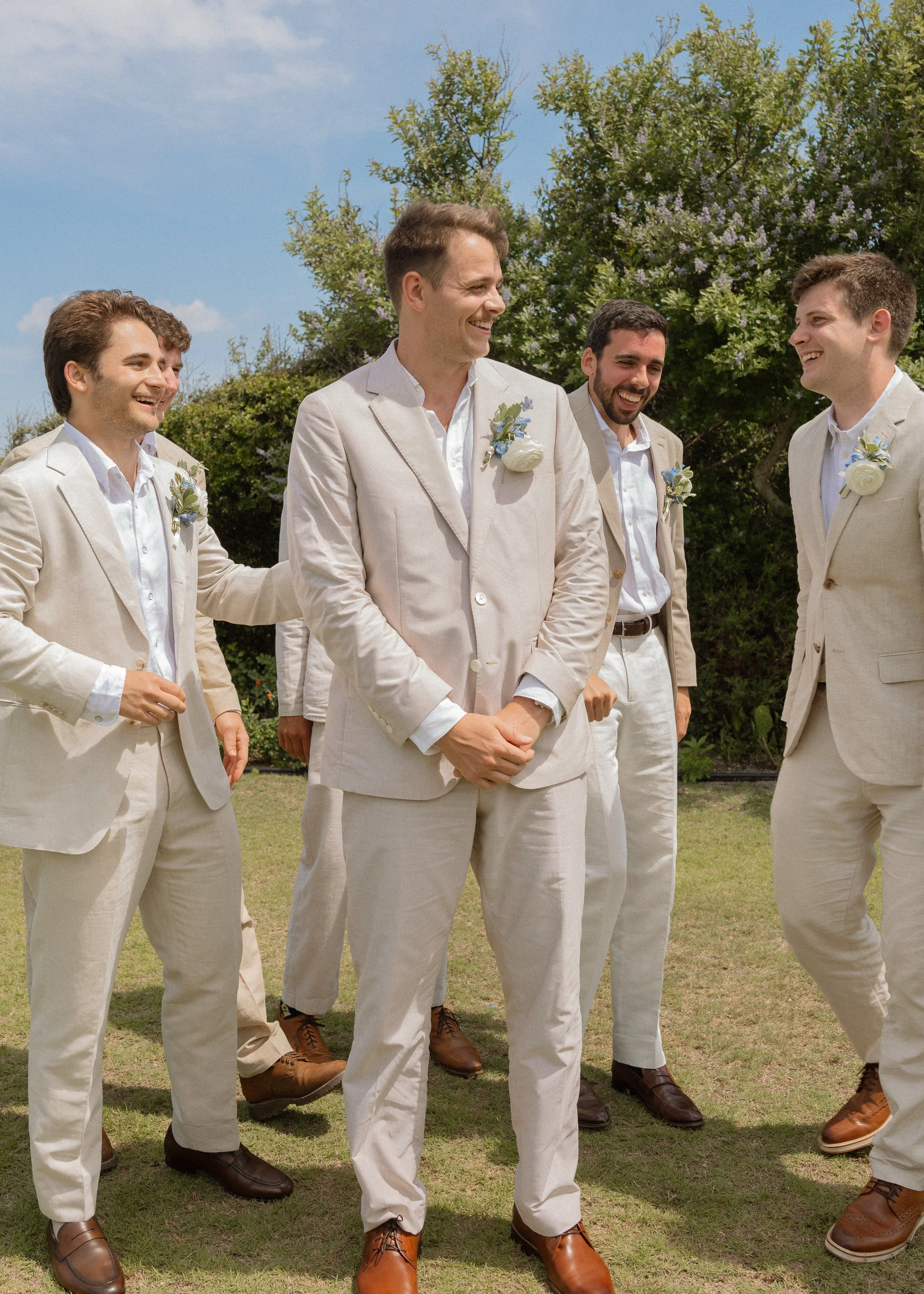 Group of five men in beige suits and white shirts outdoors, smiling and talking near bushes and trees with blue sky.