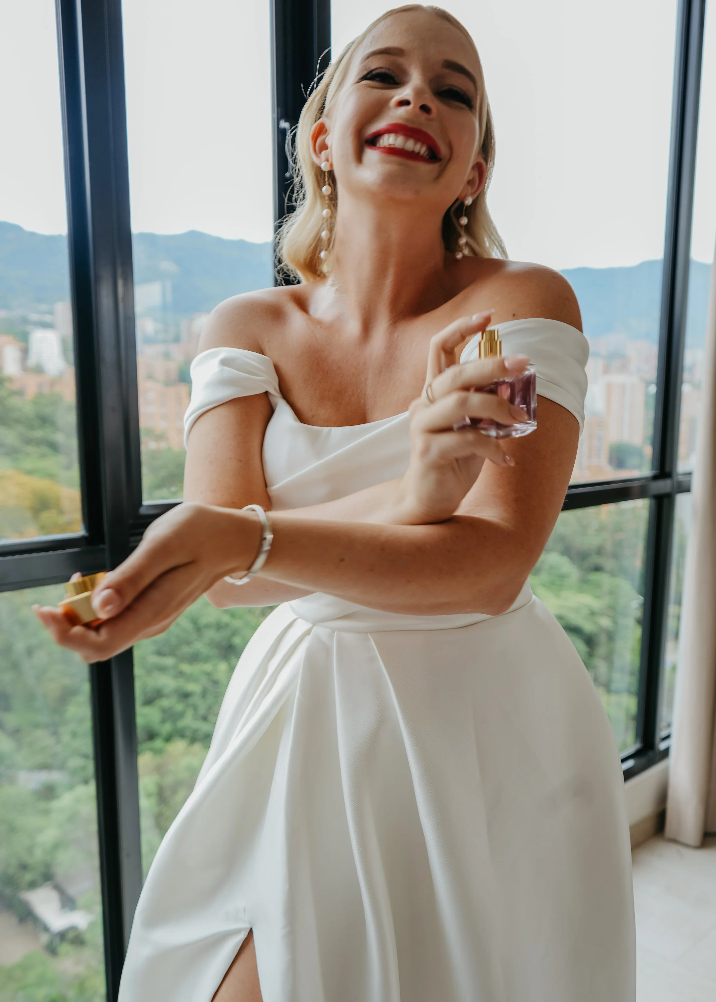 A woman in a white off-shoulder dress smiling and holding a perfume bottle in front of large windows with a city and mountain view.