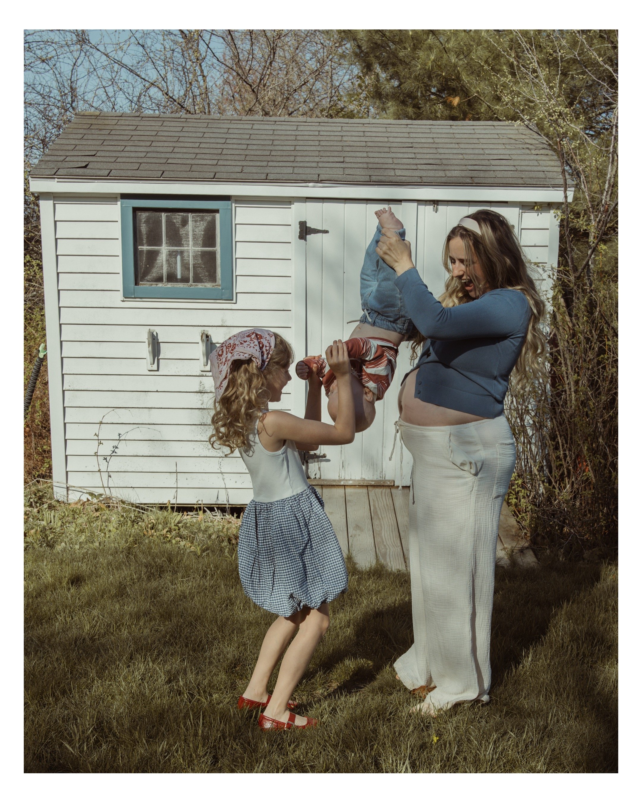 Woman lifting a girl in front of a white shed with blue trim, trees, and grass.