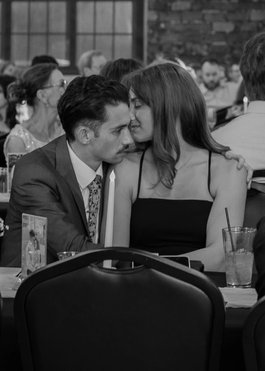 A black and white photo of a man and woman sitting closely together at a dining table during a social event. The woman is leaning her face towards the man's forehead, and they appear to be sharing an intimate moment. The table has drinks, a candle, a