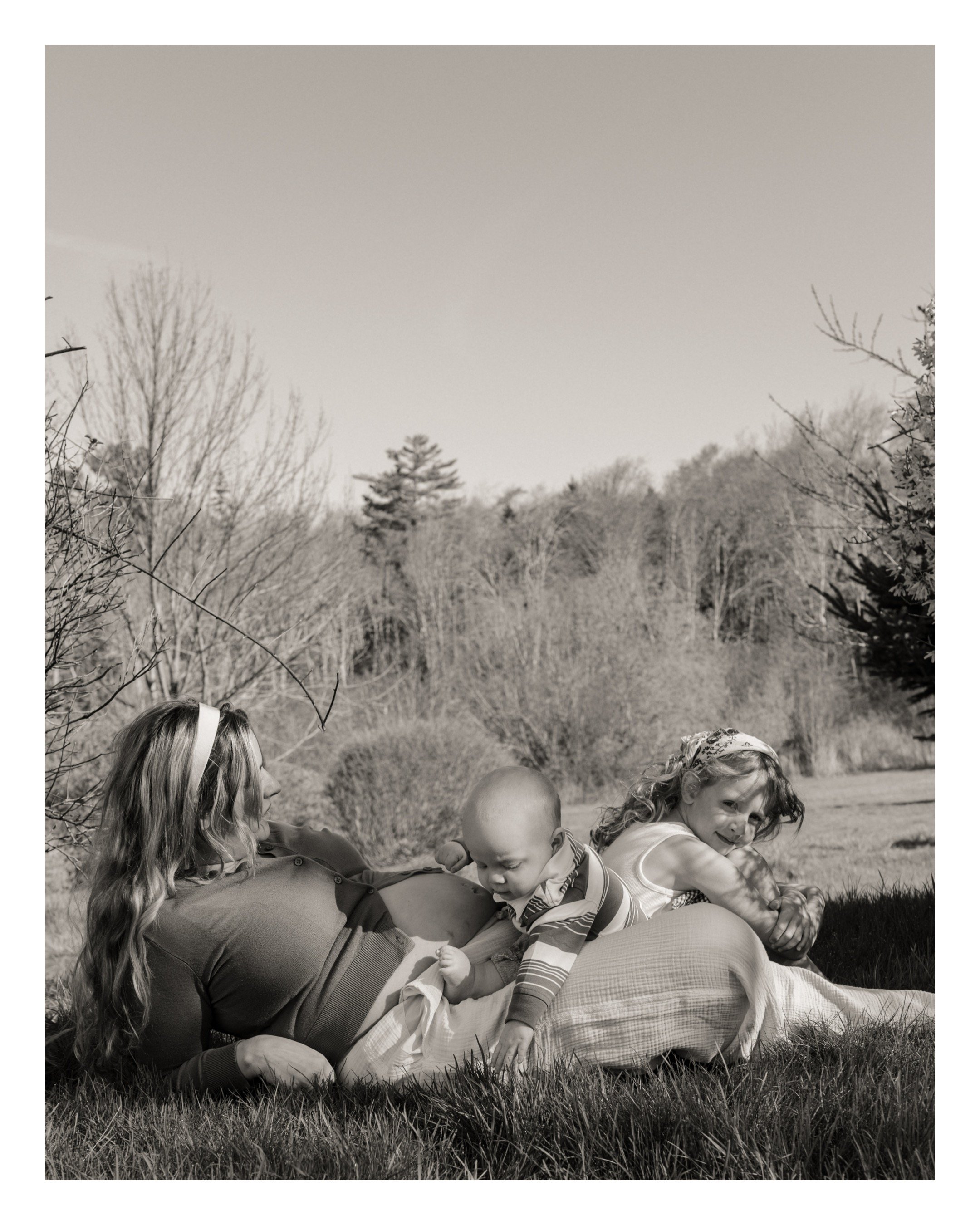 A woman with long hair lying on her side on the grass with three young children beside her, outdoors in a natural setting with trees and a cloudy sky in the background.