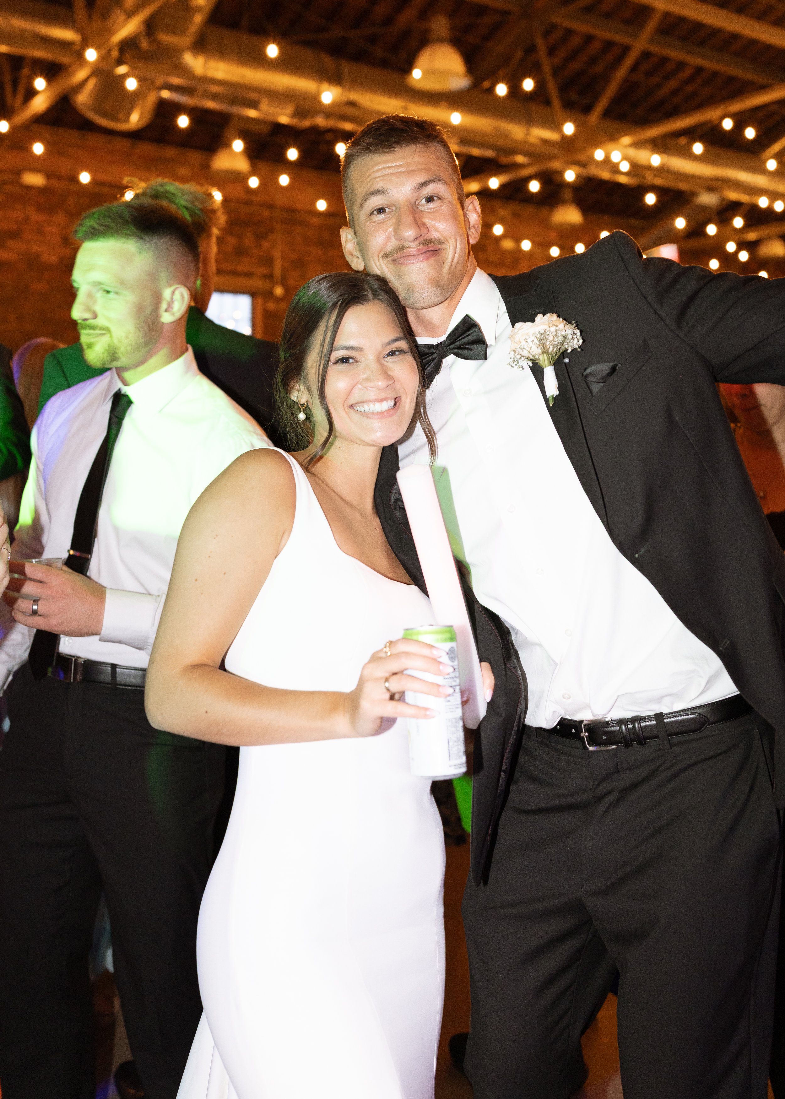 A joyful wedding celebration with a bride and groom smiling at the camera; the bride is holding a drink, and the groom is wearing a tuxedo with a boutonniere, while other guests are dressed in formal attire in a festively decorated indoor venue.