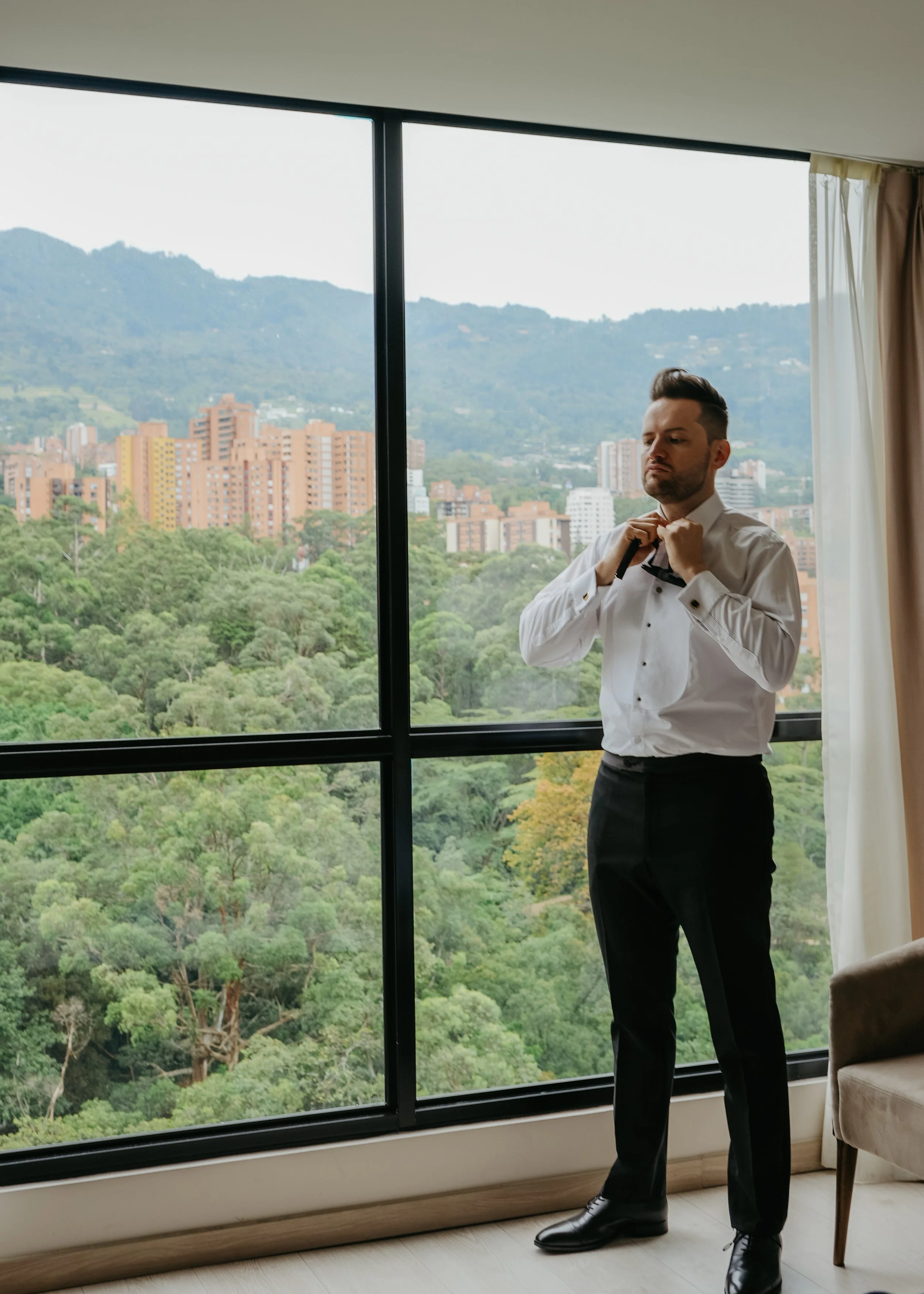 A man in a white dress shirt and black pants adjusts his black bow tie near a large window with a view of green trees and a cityscape with mountains in the background.
