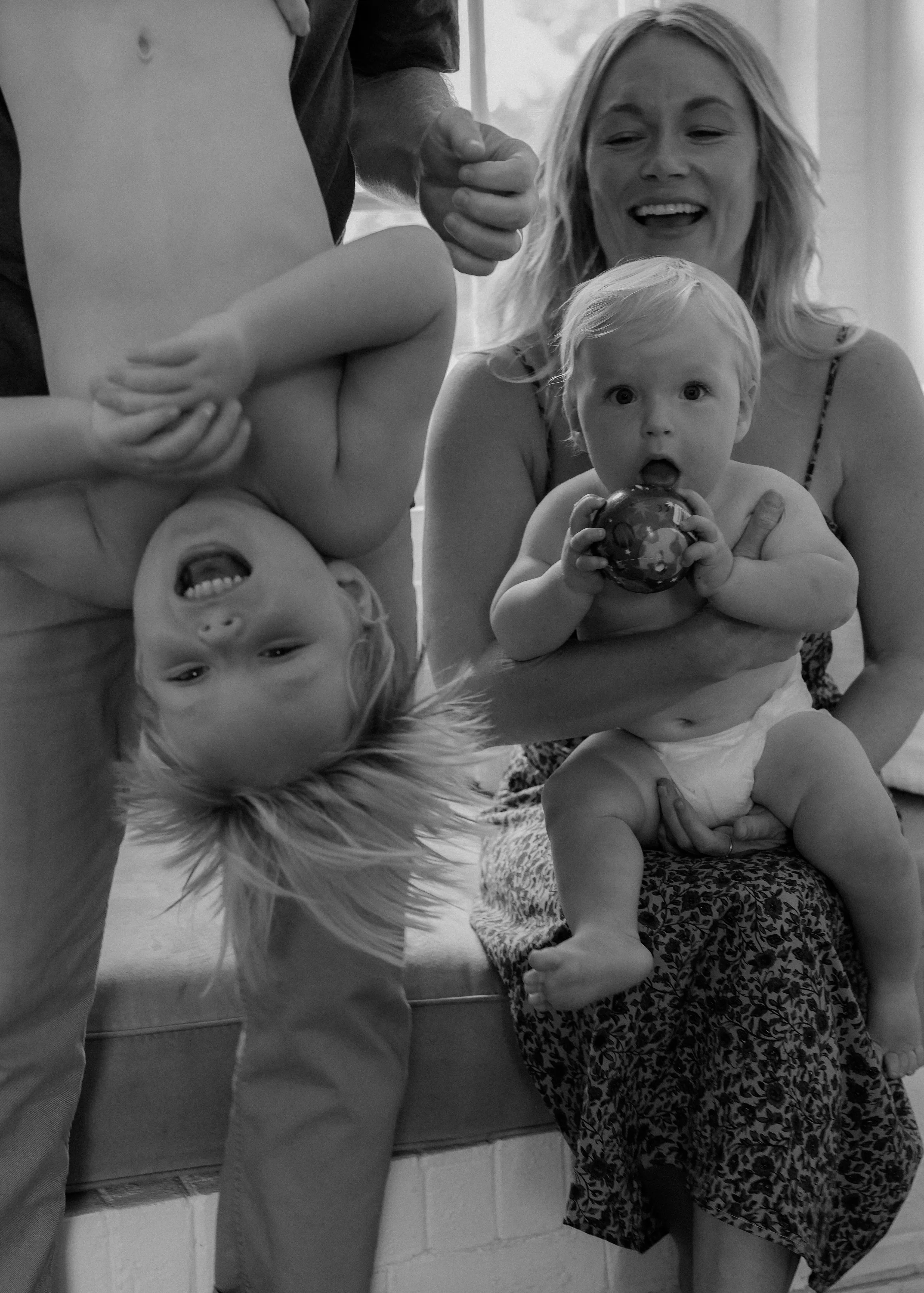 A woman holding a baby, sitting on a patterned cushion, with another baby upside-down in front of her. The babies are both holding toys; one is holding a ball, and the other's mouth is open in surprise or excitement.