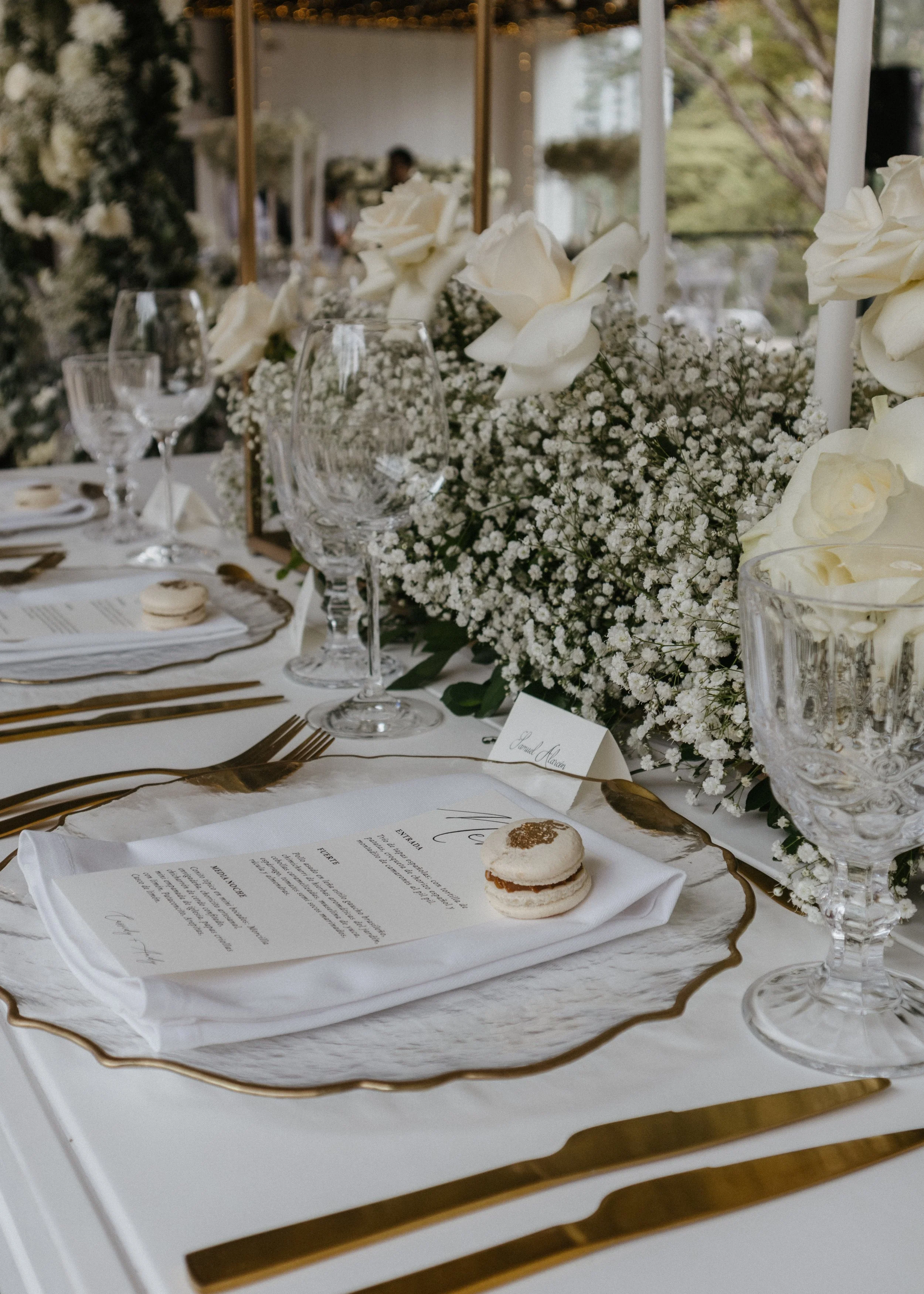 Elegant wedding reception table with white flowers, glassware, gold utensils, and a menu on a white tray, with a blurred outdoor background.