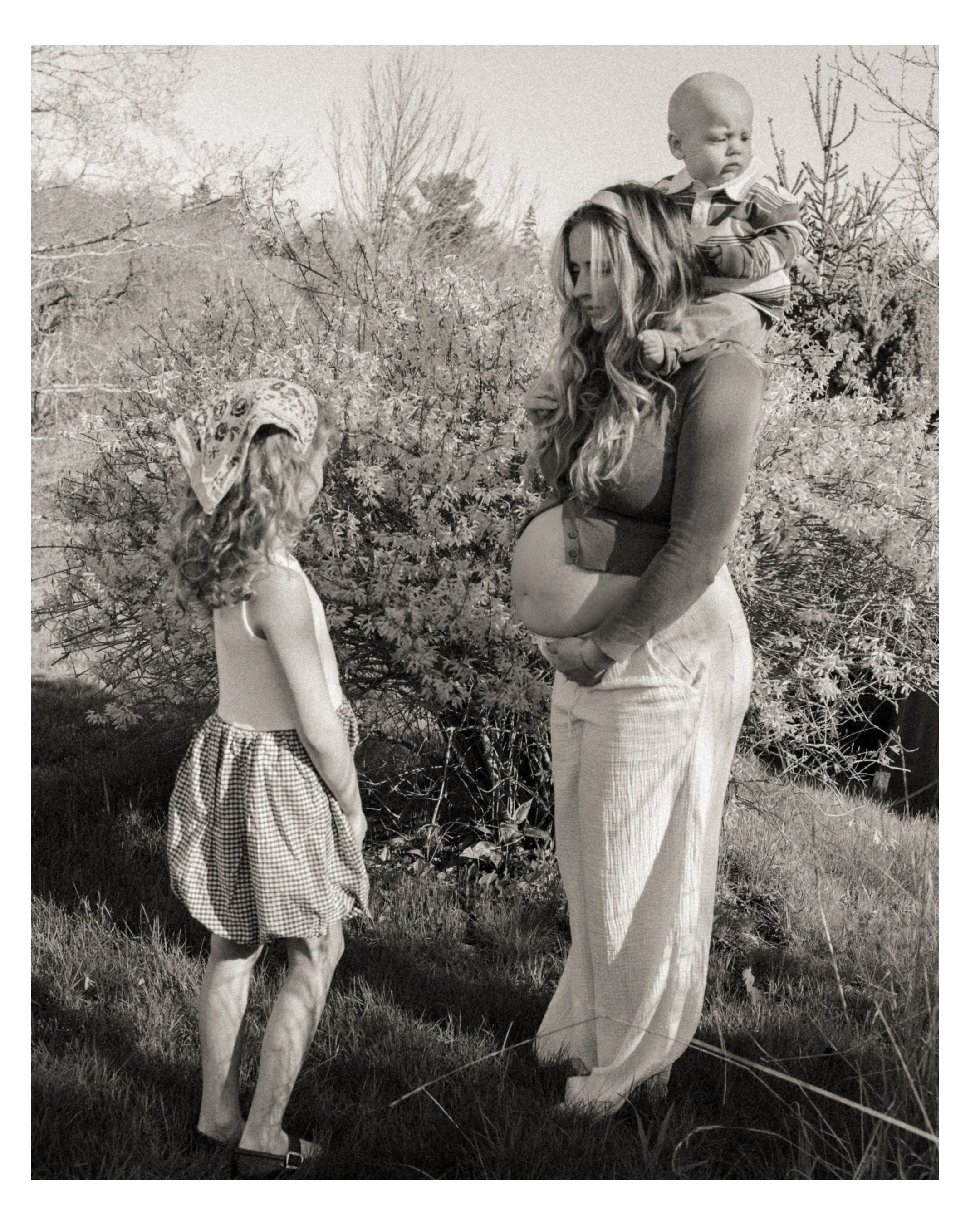 A woman with long wavy hair carrying a toddler on her back, standing outdoors among trees and bushes. The toddler has a bald head, is wearing a striped shirt, and is looking to the side. An young girl with curly hair, wearing a checkered dress and a 