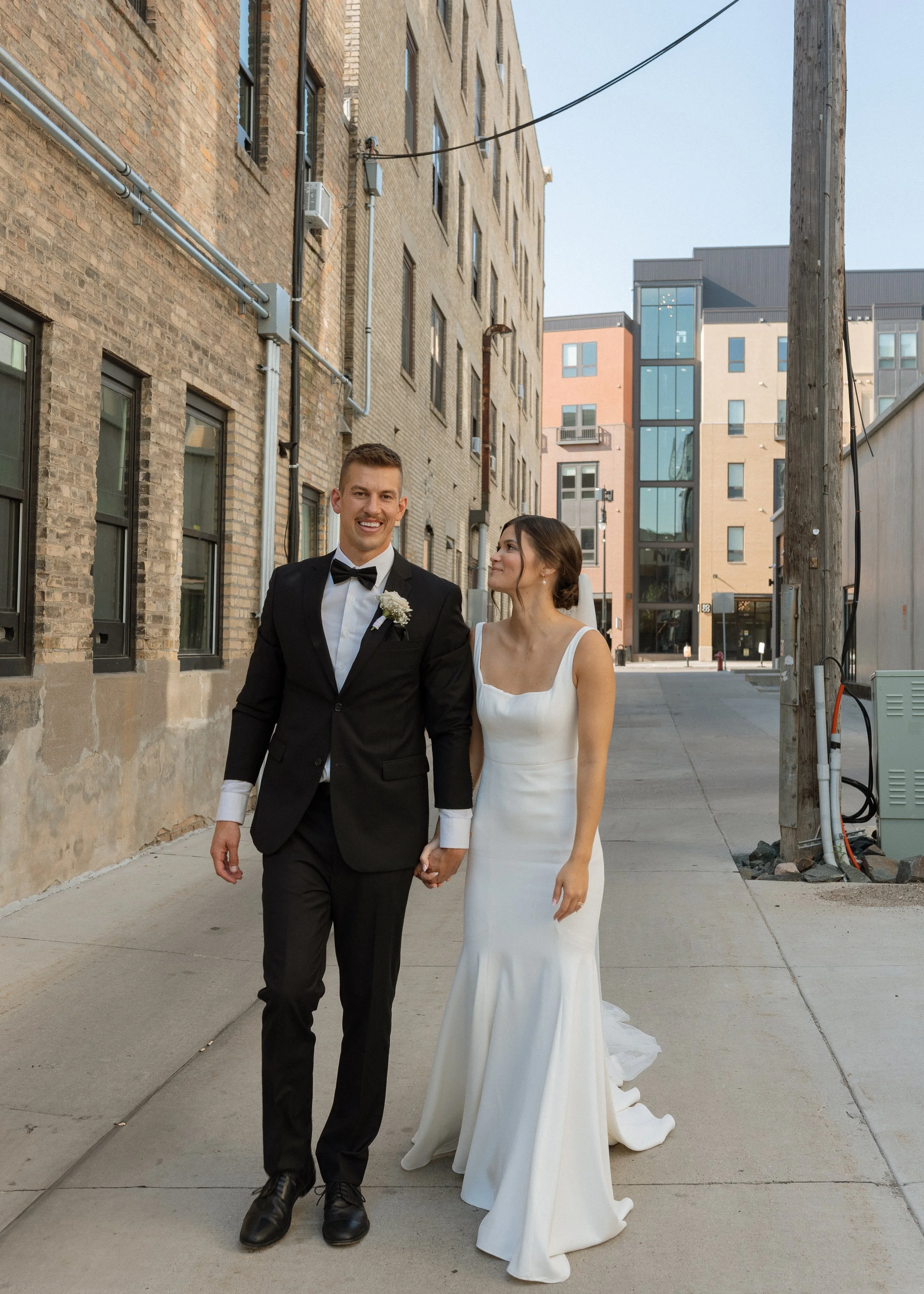 A newlywed couple walking hand in hand on an urban sidewalk, with brick and modern buildings in the background. The groom wears a black tuxedo with a bow tie, and the bride wears a white wedding gown with a train.
