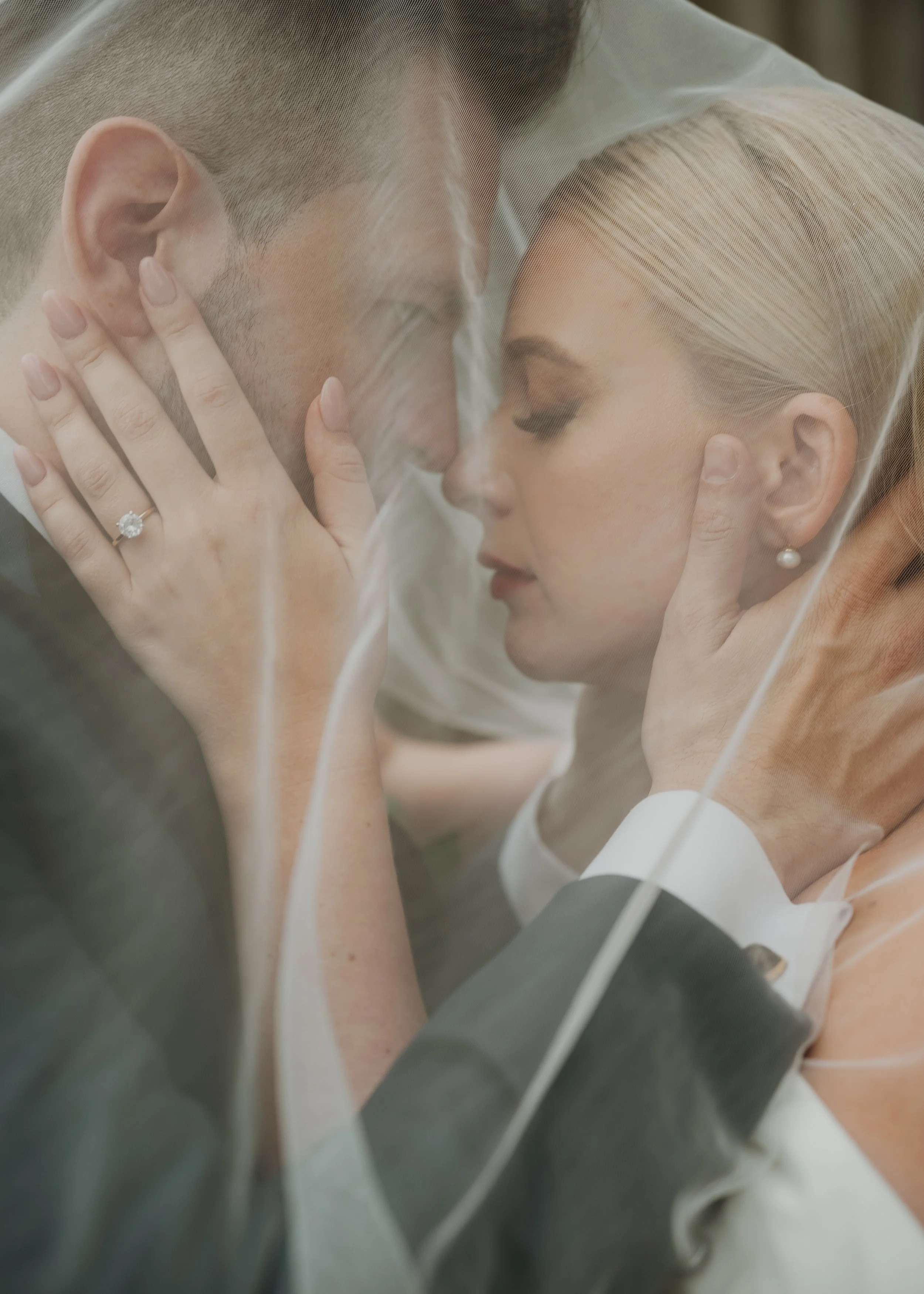 A bride and groom kiss behind a wedding veil, with the bride wearing pearl earrings and a ring, and the groom with a short haircut.