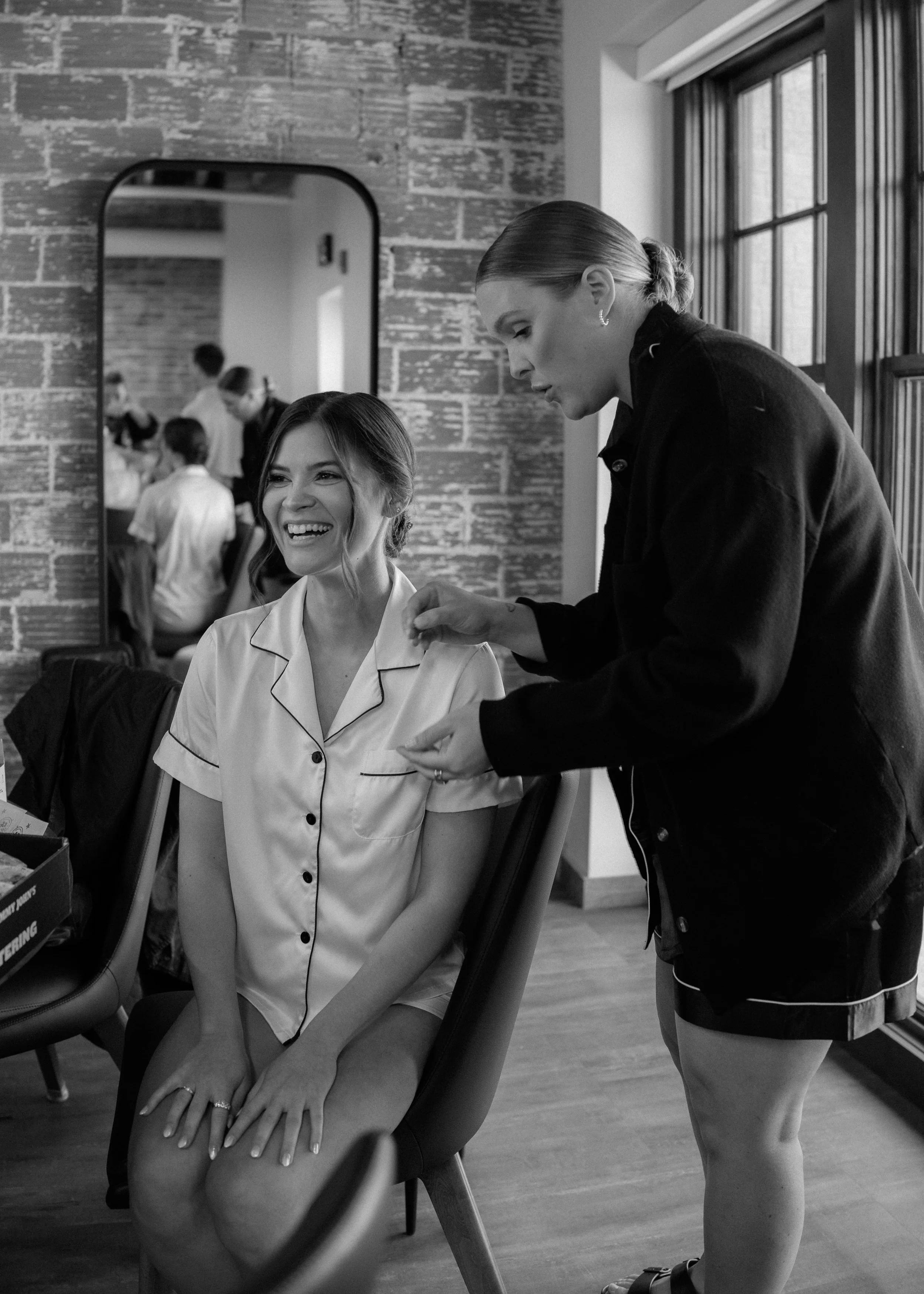 A woman sitting in a chair, smiling, wearing a satin pajama top, with her hands resting on her lap, as another woman adjusts her appearance, possibly a makeup artist or hairstylist, in a room with large windows and a brick wall.