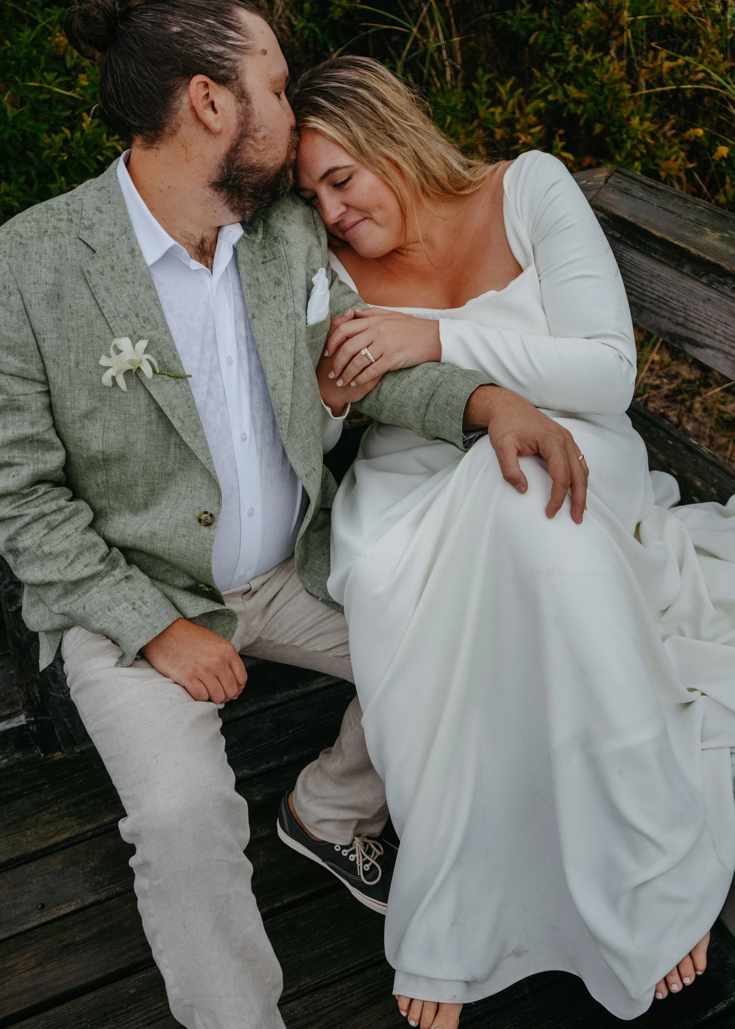 A man and woman sitting close together on a wooden bench outdoors, smiling and resting their heads against each other, with their eyes closed. The man is wearing a gray blazer, white shirt, and beige pants with a white flower on his lapel. The woman 