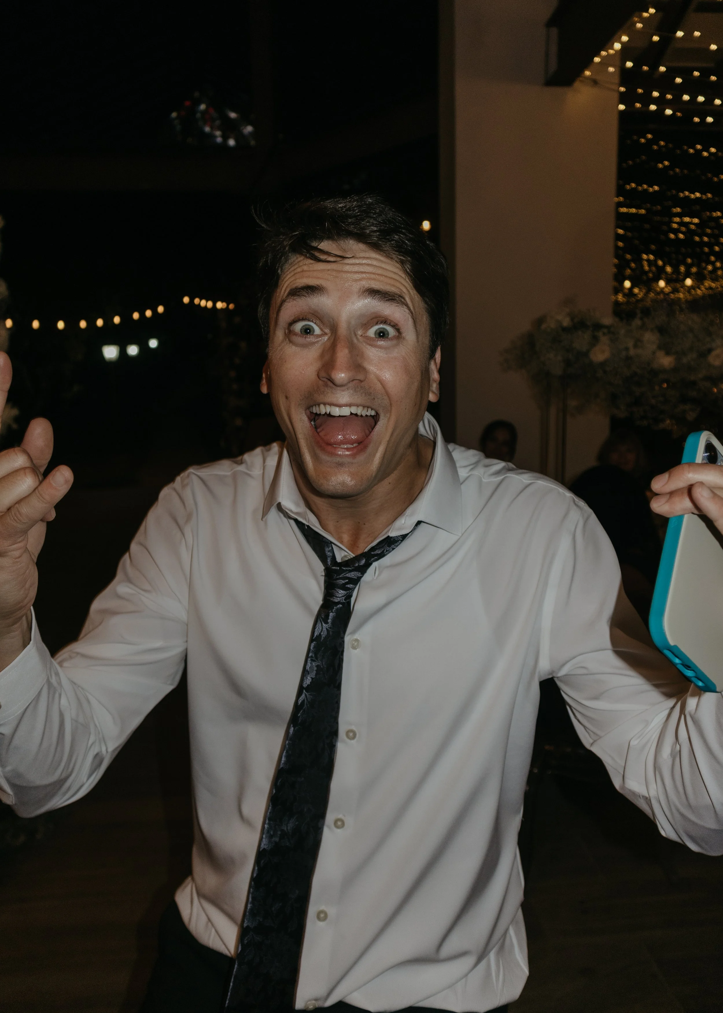 A man with dark hair and bright eyes, wearing a white shirt and a black patterned tie, excitedly smiling with his mouth open and thumbs up at an indoor party with string lights and other people in the background.