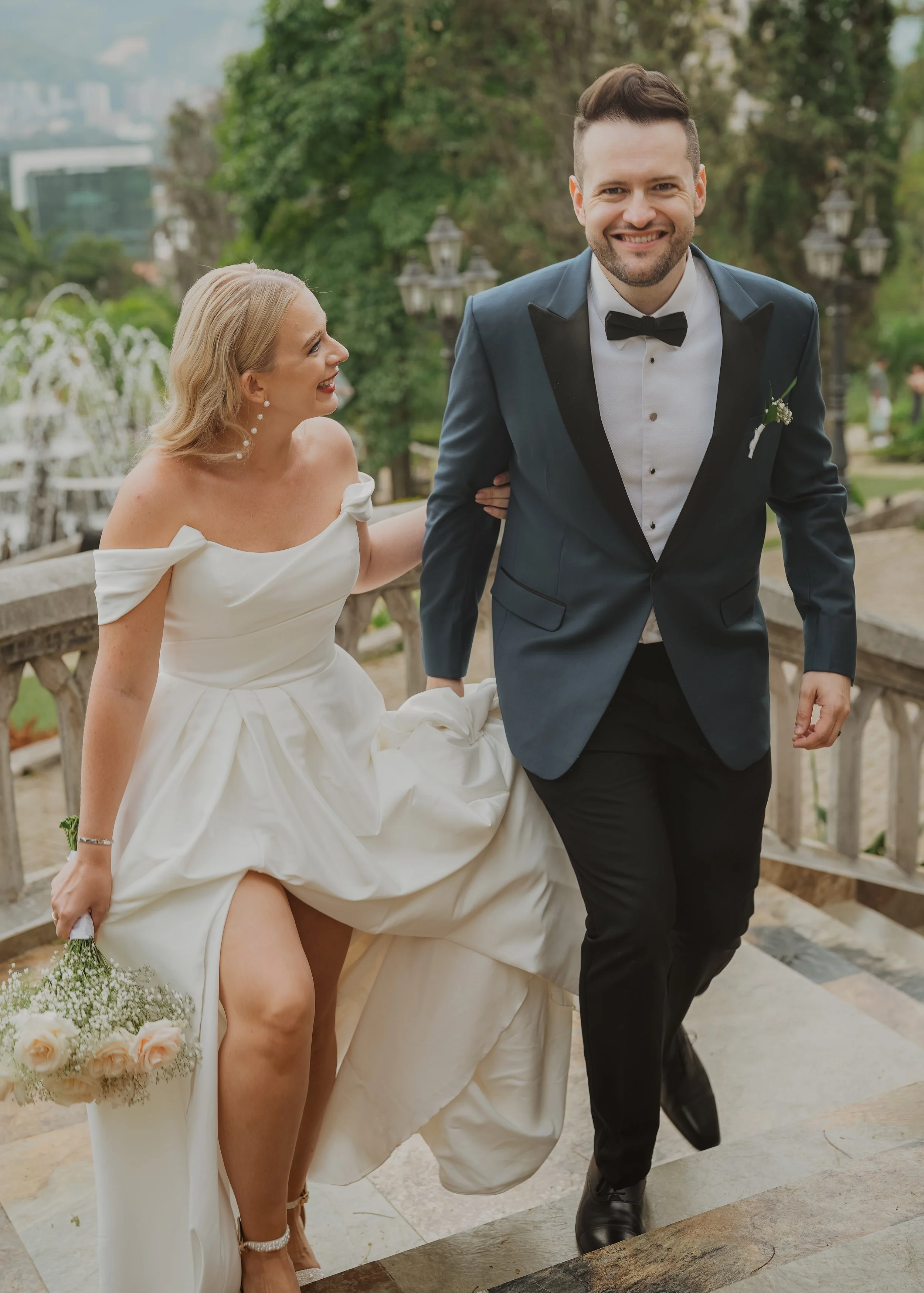 A bride and groom walking down stairs outdoors, smiling. The bride is in a white wedding dress holding a bouquet, and the groom is in a navy tuxedo with a black bow tie.
