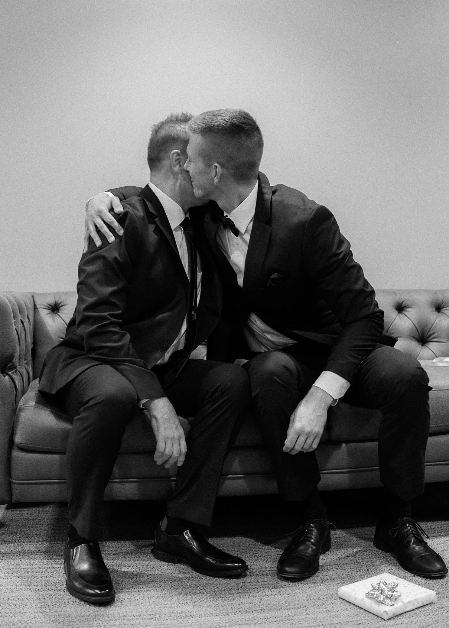 Two men in suits sitting close on a couch, kissing each other.