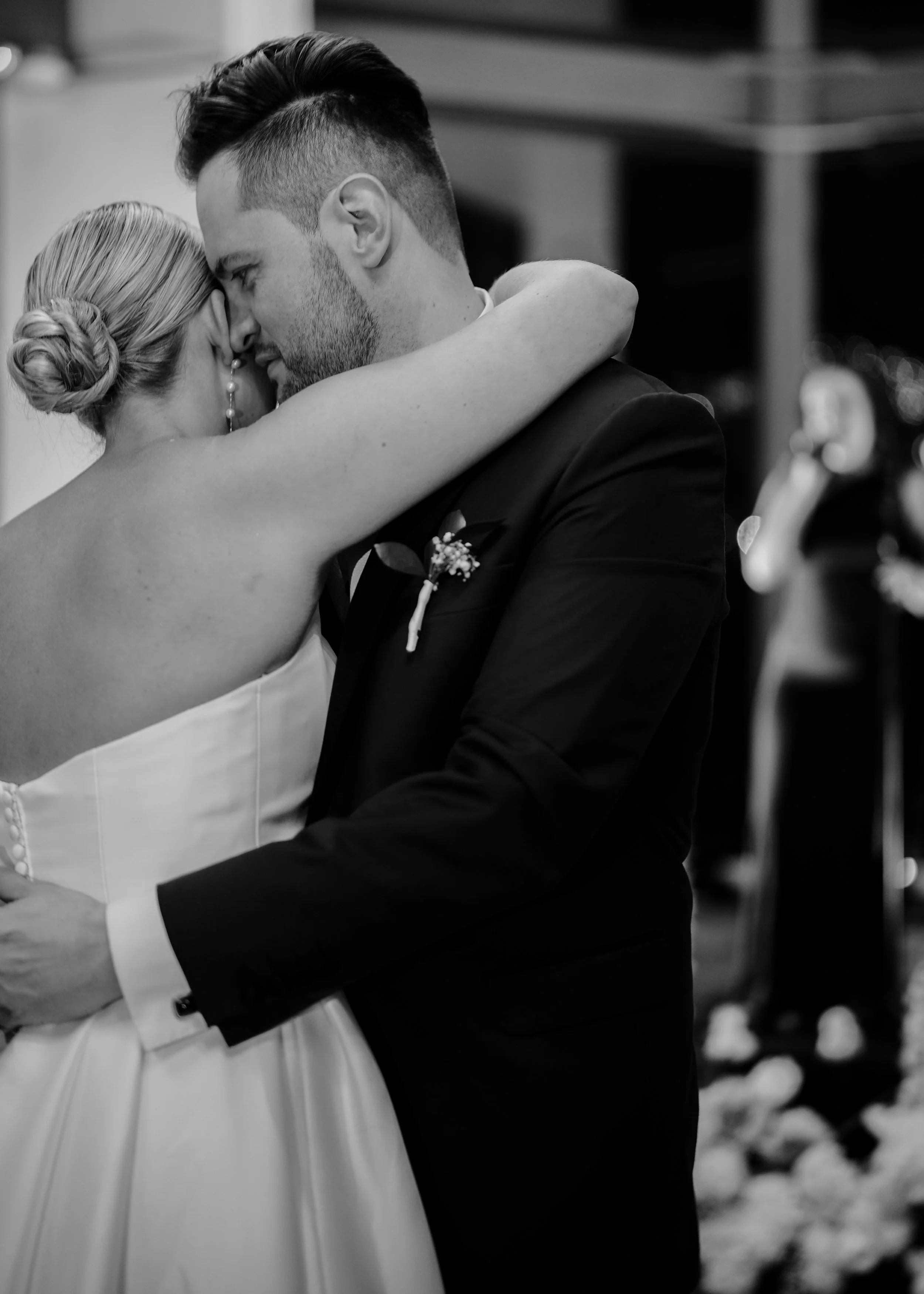 A black-and-white photo of a couple dancing closely at a wedding, with the woman wearing a wedding dress and the man in a suit, holding each other with their foreheads touching.