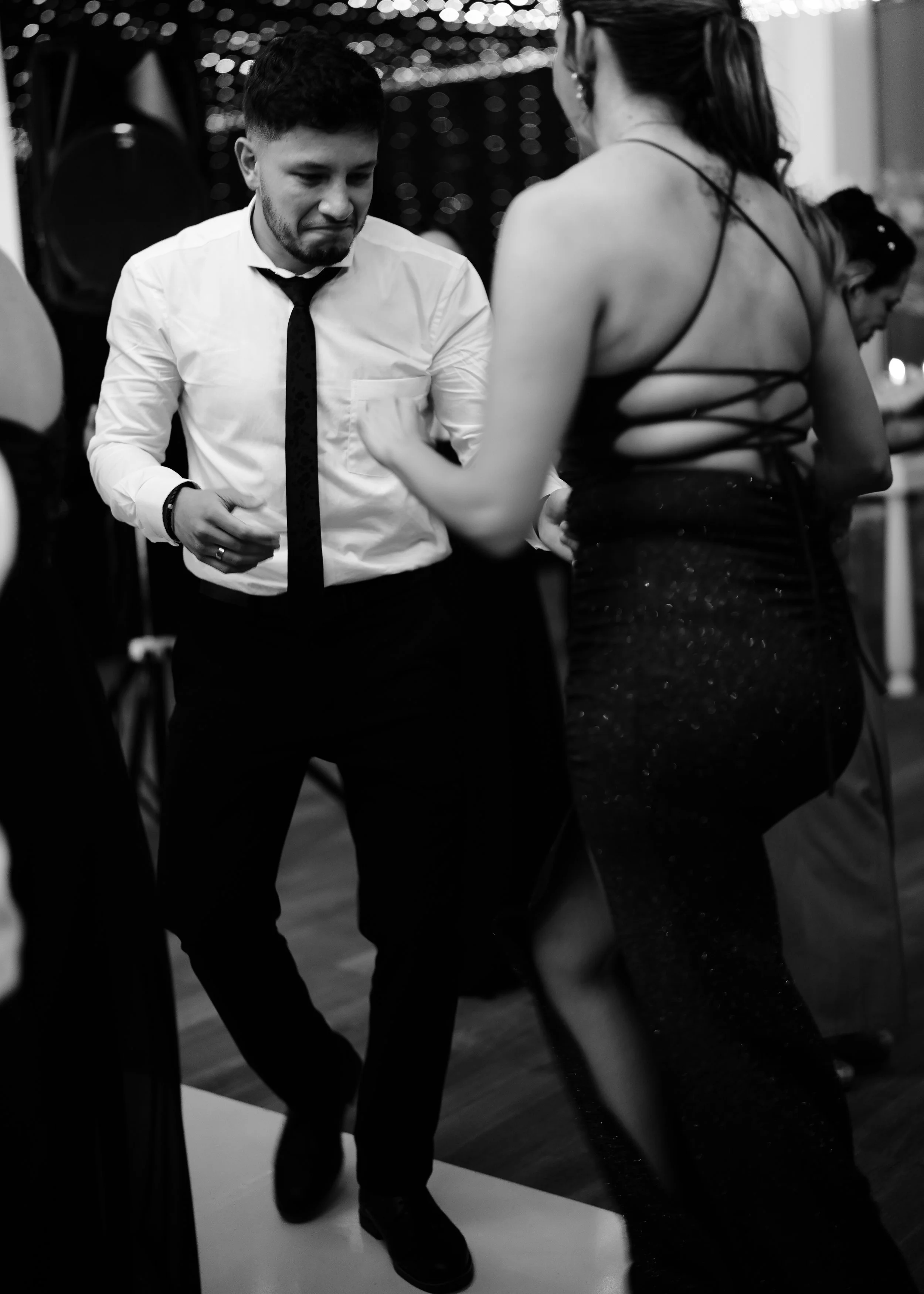 A black and white photo of a man and woman dancing at a social event. The man is wearing a white shirt, black tie, and black pants, and the woman is wearing a form-fitting dress with strappy back details. The background shows other people and a dark,
