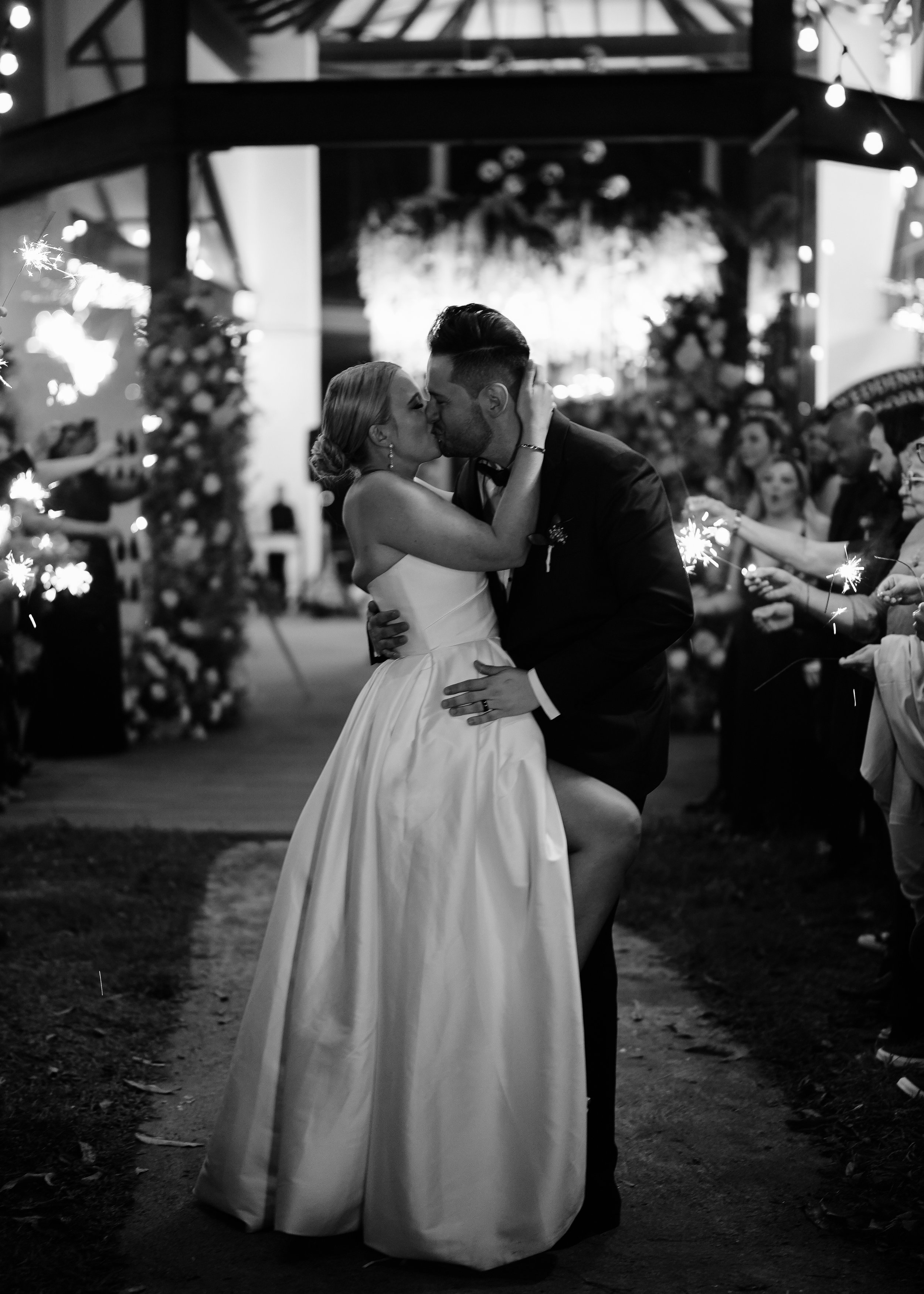 Black and white photo of a bride and groom sharing a kiss during their wedding reception, surrounded by guests holding sparklers, with a decorated backdrop and string lights overhead.