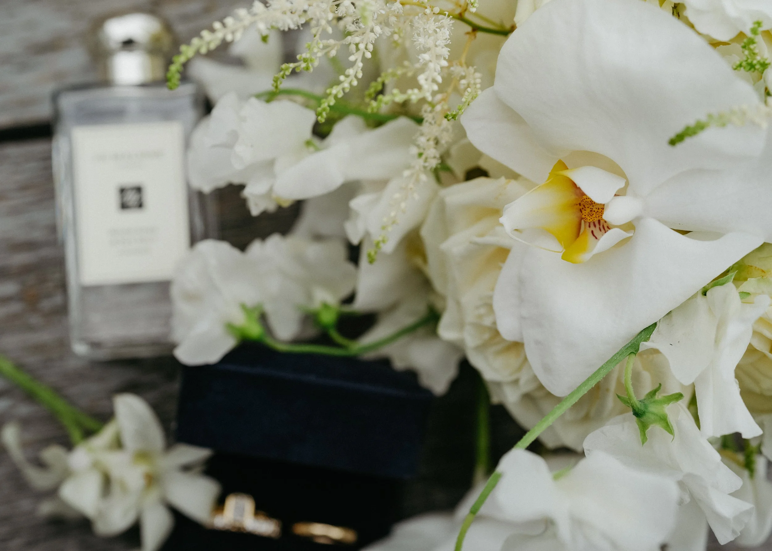 Close-up of white orchids and small white flowers on a wooden surface with a blurred perfume bottle in the background.