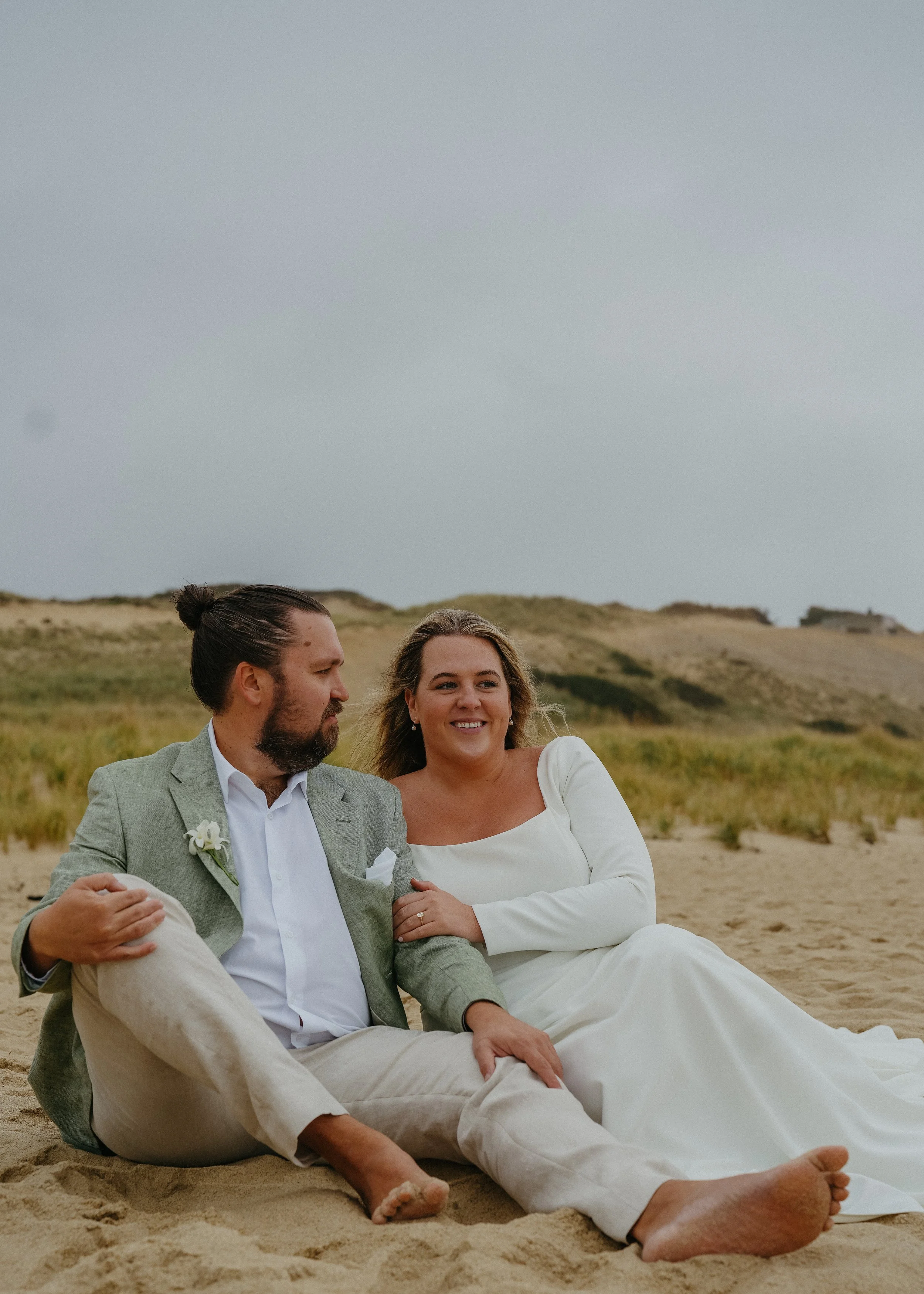 A married couple sitting on the beach, dressed in wedding clothing, smiling and enjoying the moment.