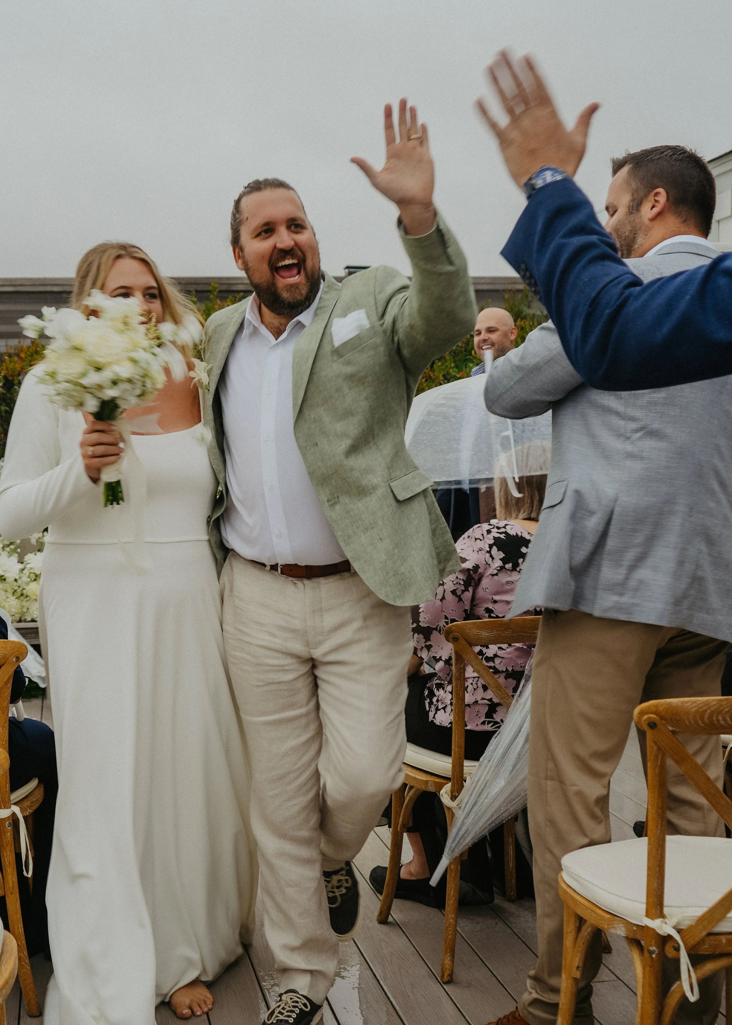 A group of people celebrating at a wedding, including a woman in a white dress holding a bouquet and a man in a light green jacket giving a high five to another man in a blue jacket.