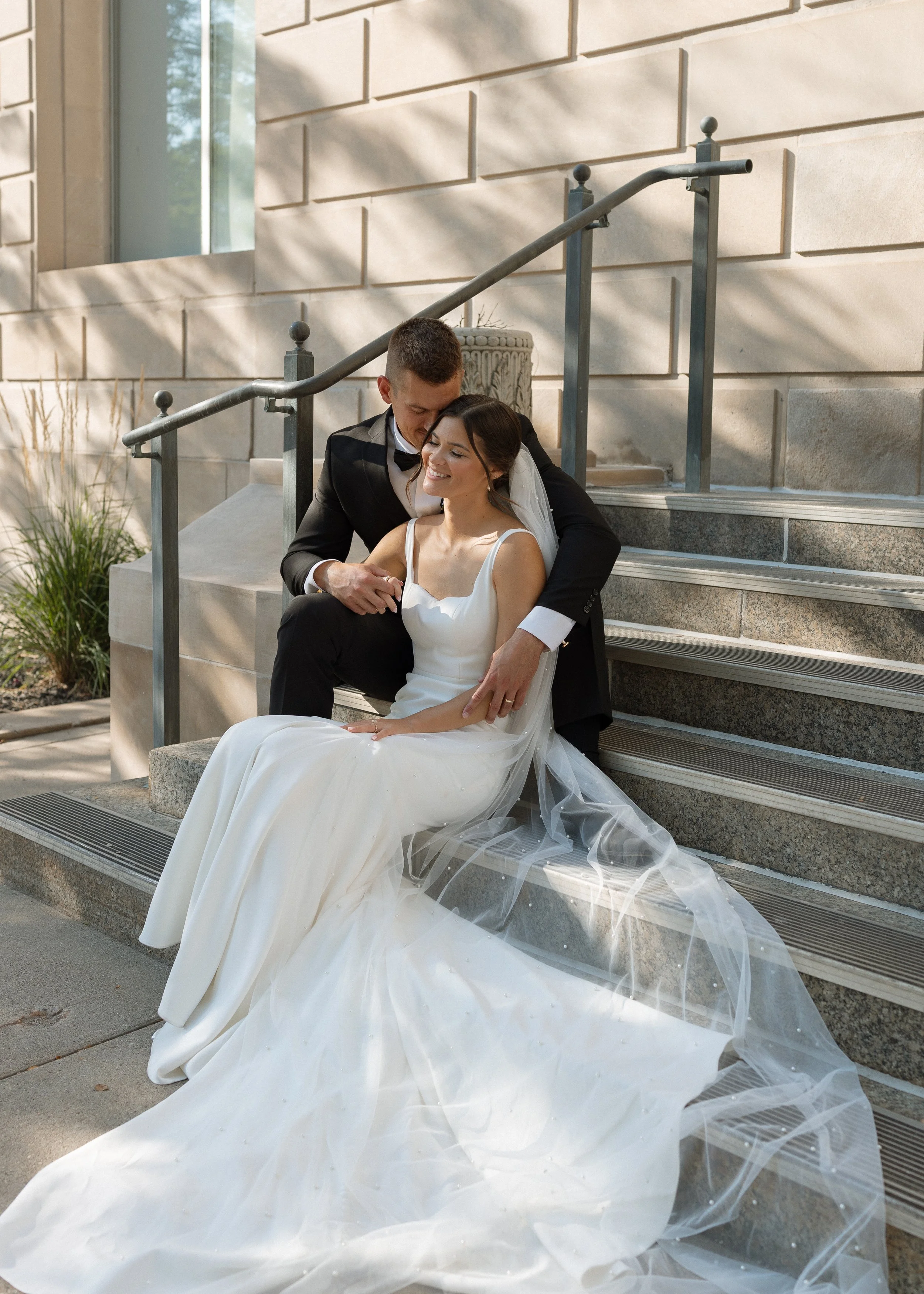 A newlywed couple sitting on outdoor stairs, with the groom in a black tuxedo and the bride in a white wedding gown, sharing a tender moment amid soft sunlight and shadows on the wall behind them.