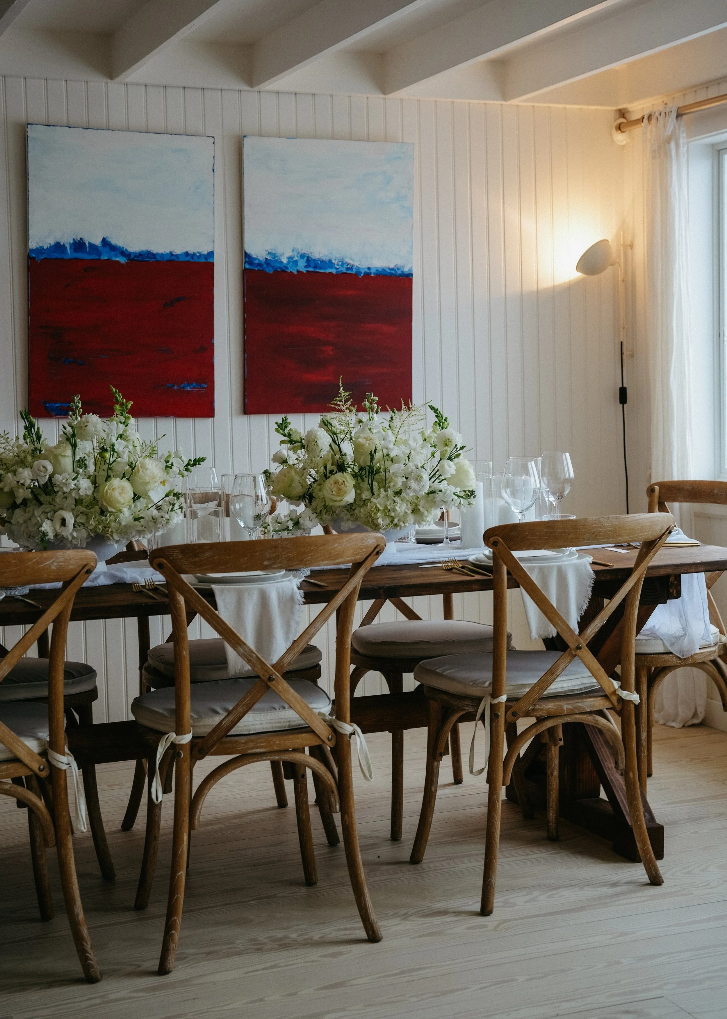 A dining room with a wooden table set with white plates, silverware, and wine glasses. A large floral centerpiece with white flowers decorates the table. Surrounding the table are wooden chairs with cushioned seats. The wall behind features two abstr