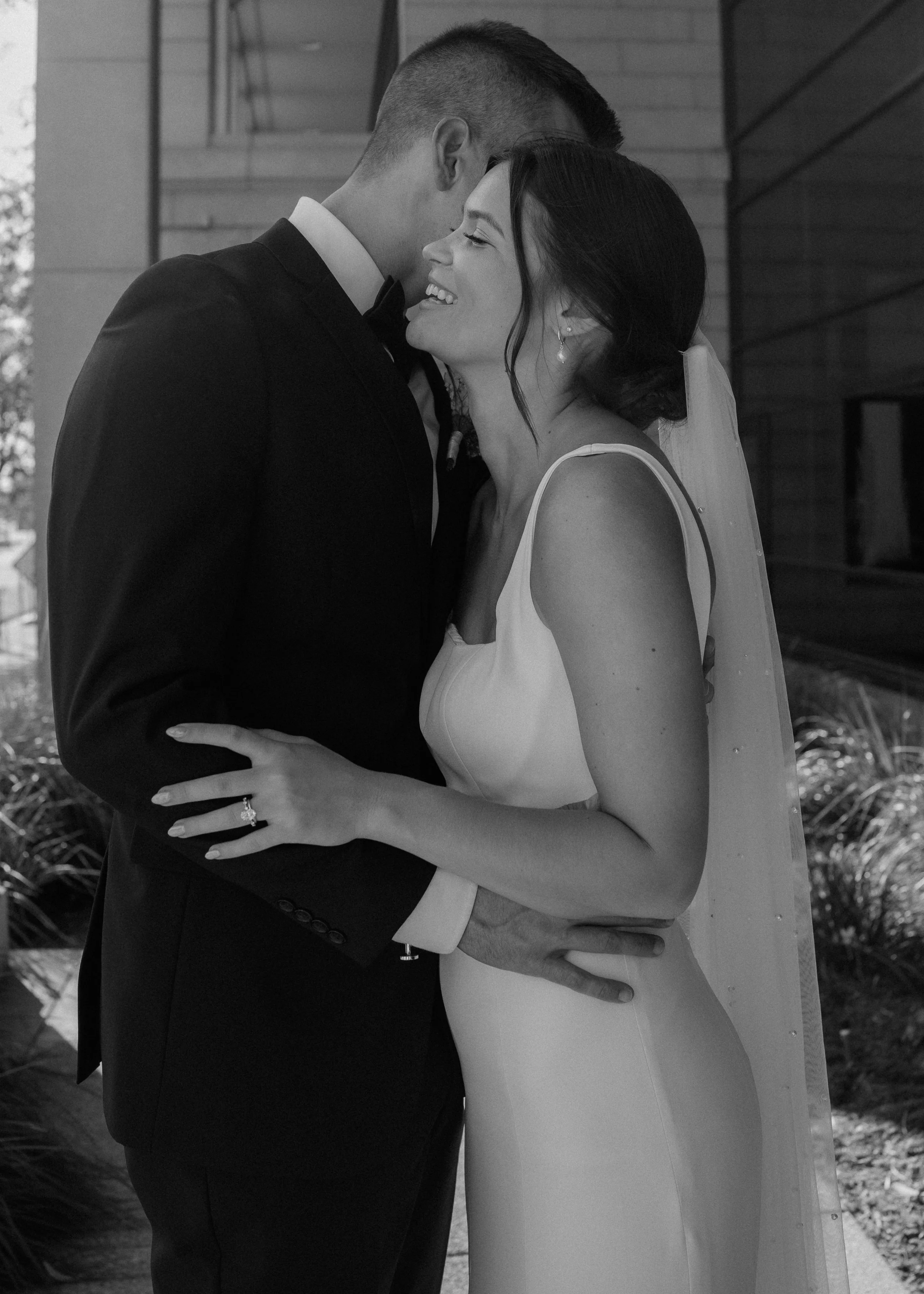 A black-and-white photo of a bride and groom embracing on their wedding day, smiling and standing close together. The groom is in a tuxedo, and the bride is wearing a wedding dress and veil.