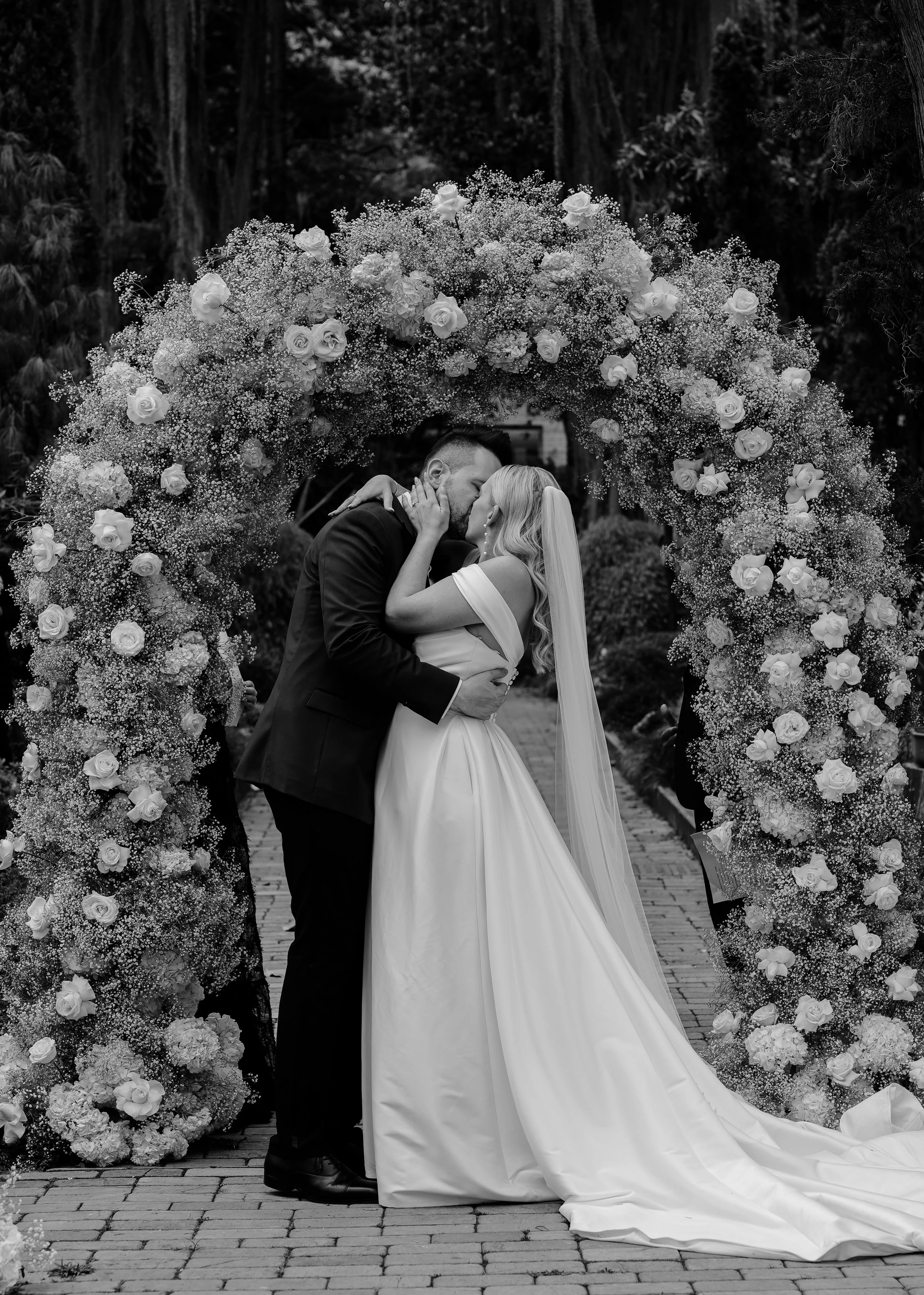 A black and white photo of a bride and groom kissing under a floral arch at their wedding. The arch is decorated with roses and baby's breath, and the couple is standing on a brick pathway in an outdoor garden setting.