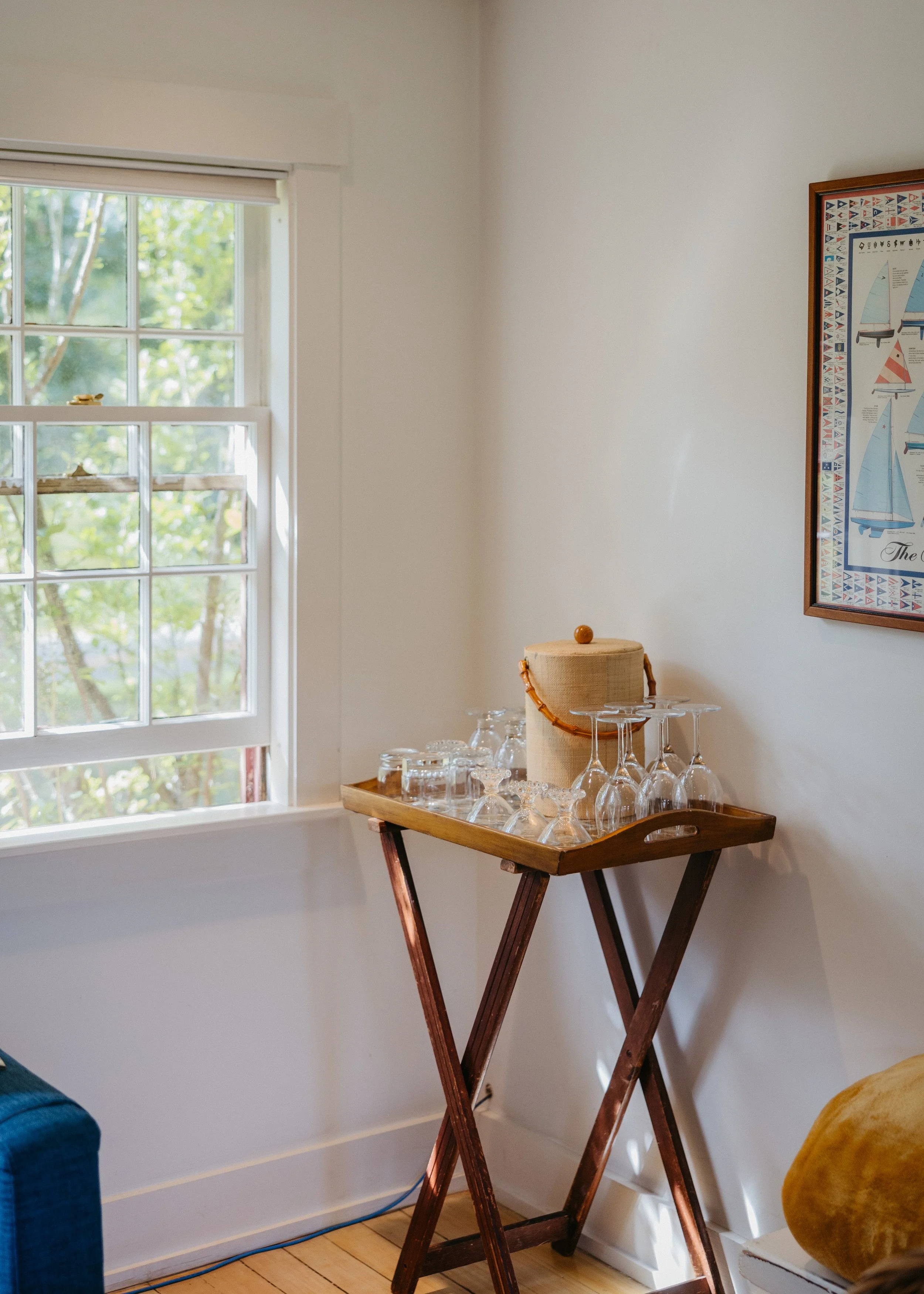 A wooden serving tray with upside-down wine glasses and a beige fabric container on a foldable wooden table in a bright corner of a room. A window with multiple panes and white trim shows green trees outside. A framed nautical chart hangs on the white wall.
